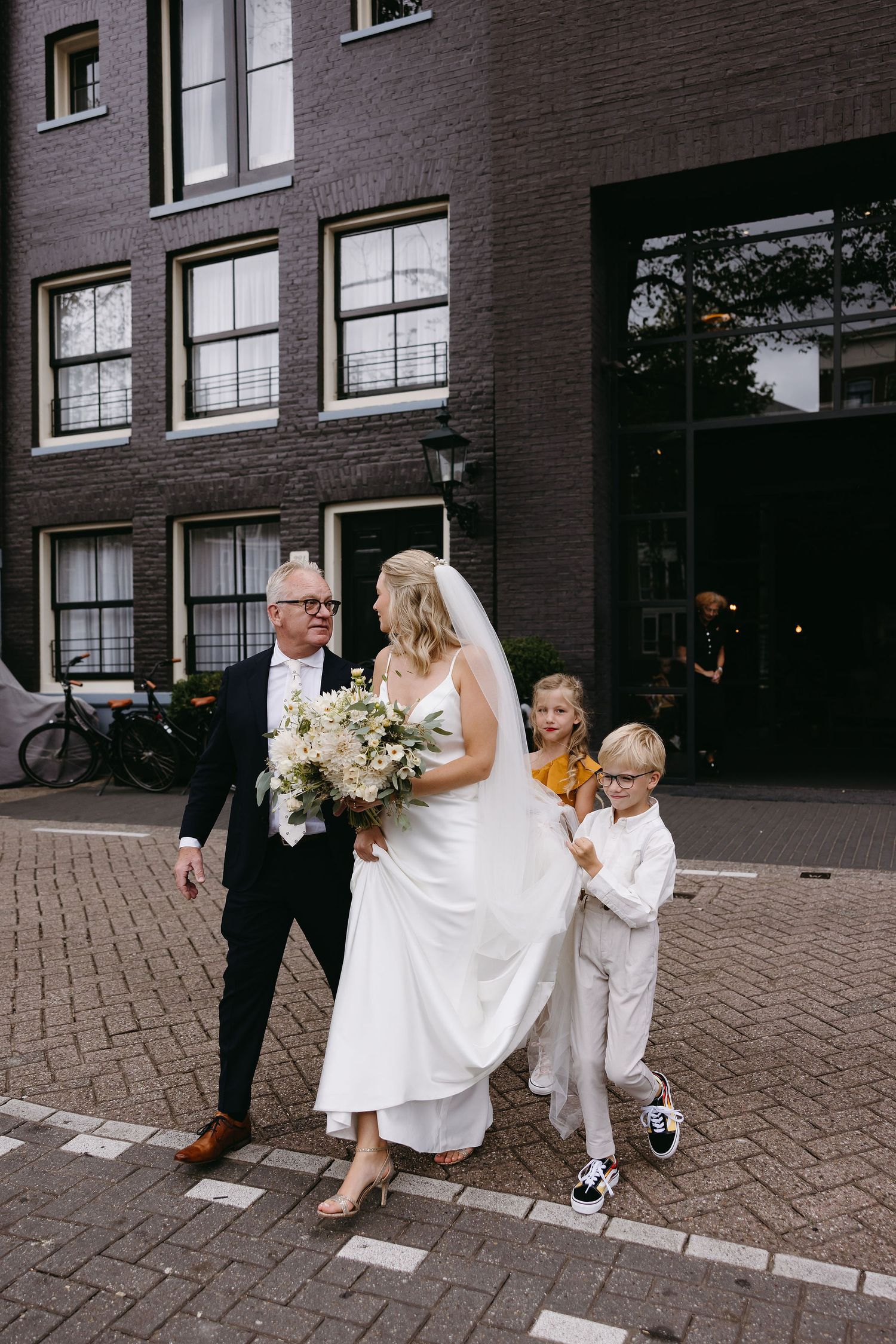 Bride holding bouquet walks with guests on cobblestone street.