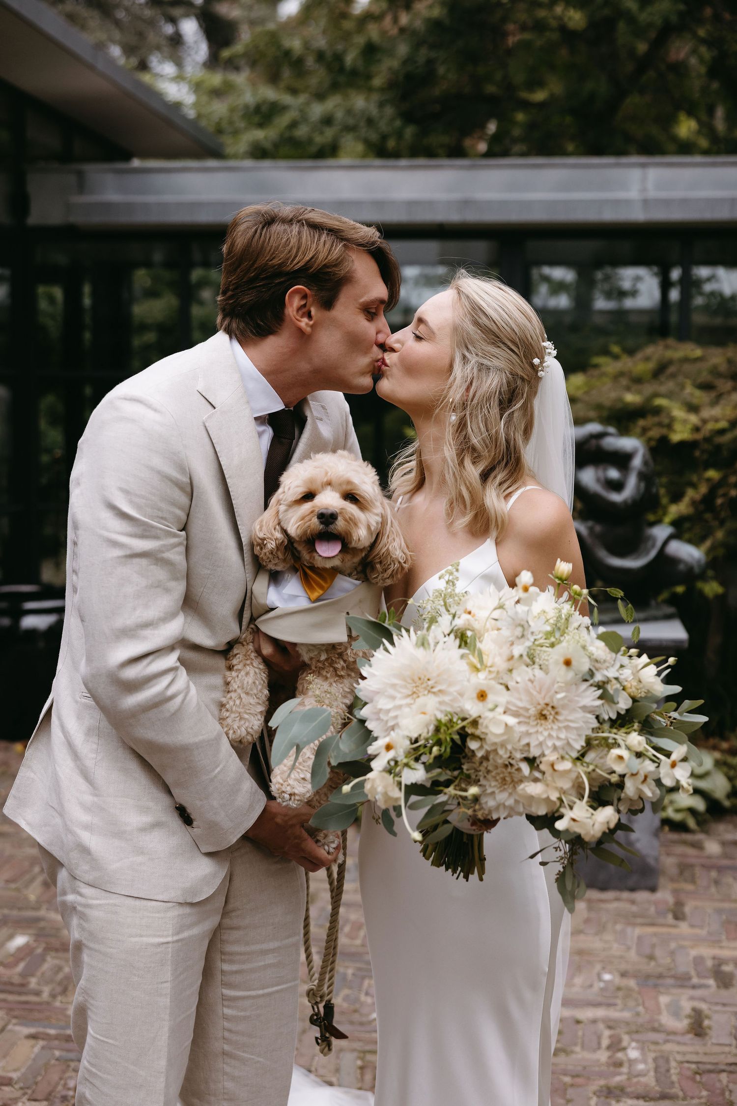 Couple in white attire sharing intimate moment while holding small dog and floral bouquet.