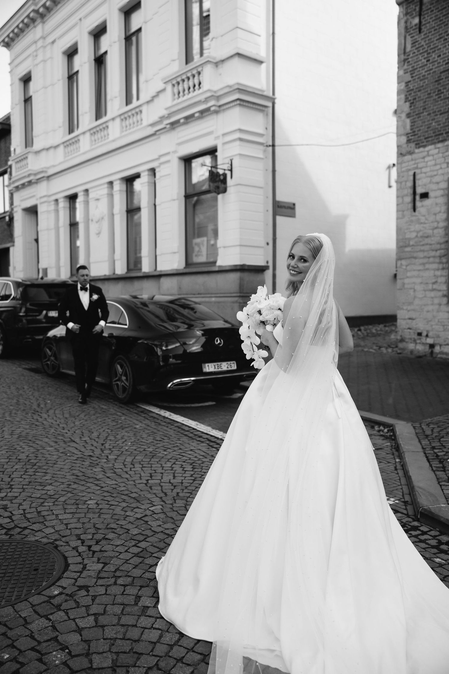 A bride in flowing white dress and veil stands on cobblestone street near classic car.