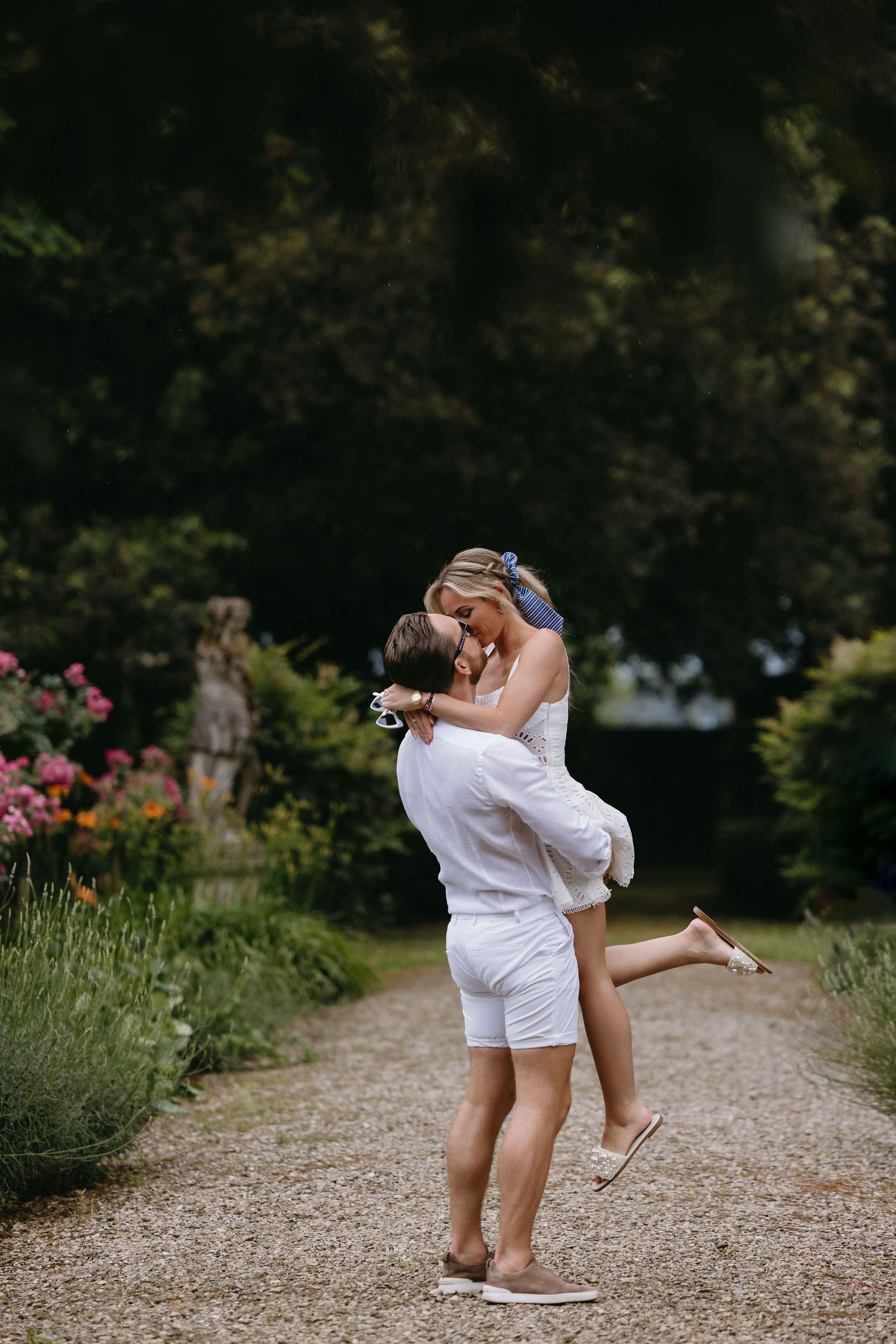 Two people in white outfits sharing a joyful moment on a garden path surrounded by flowers and greenery.