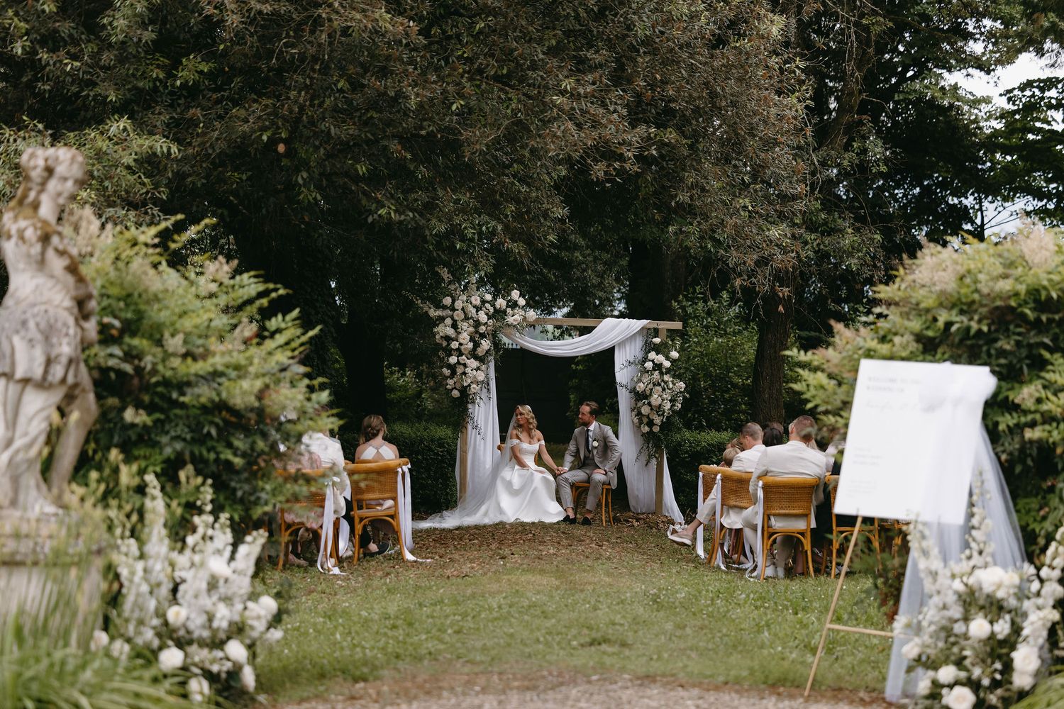 Intimate outdoor garden wedding ceremony setup with white chairs and floral arch under trees.