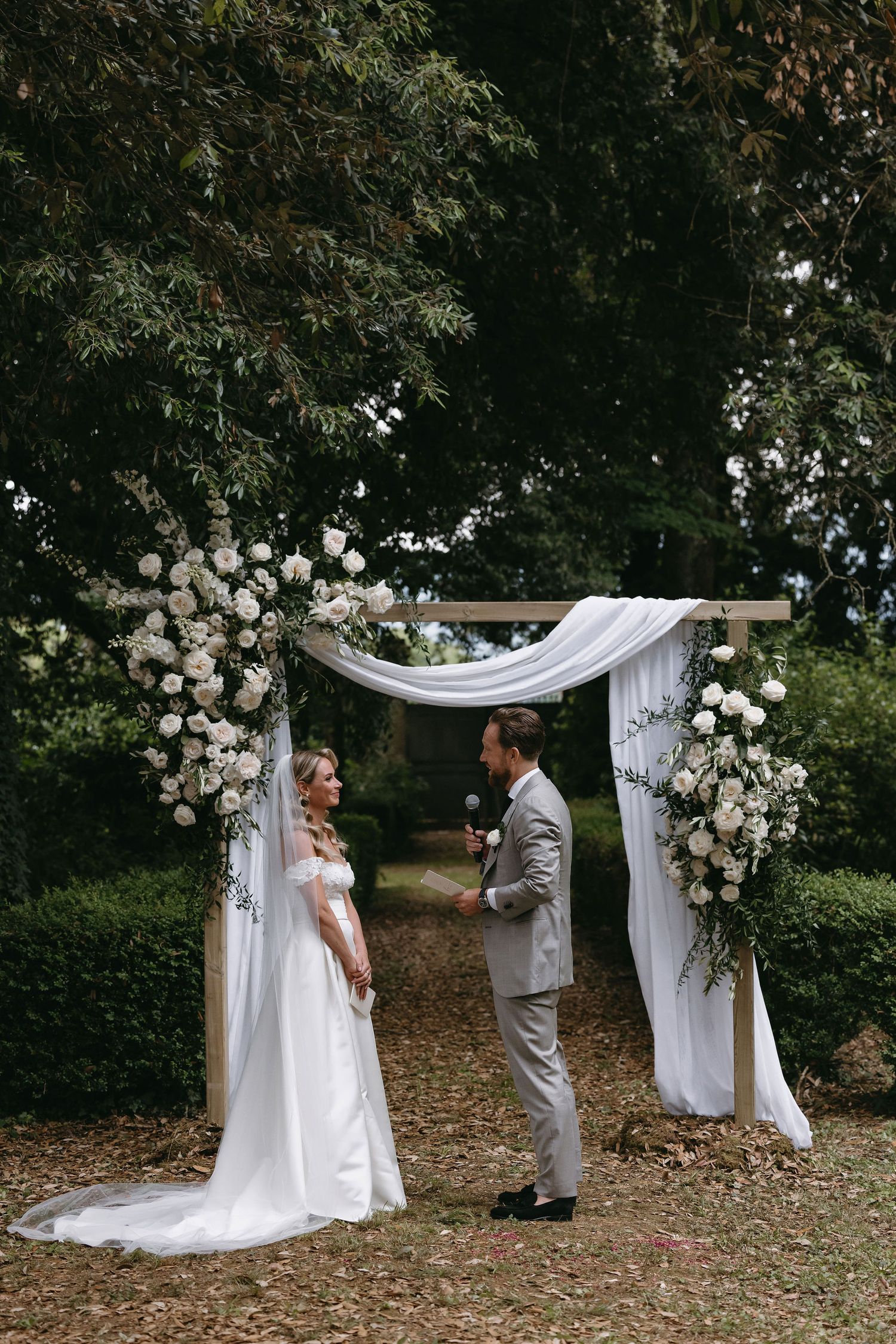 Wedding ceremony setup with white floral arch and draped fabric in a garden setting.