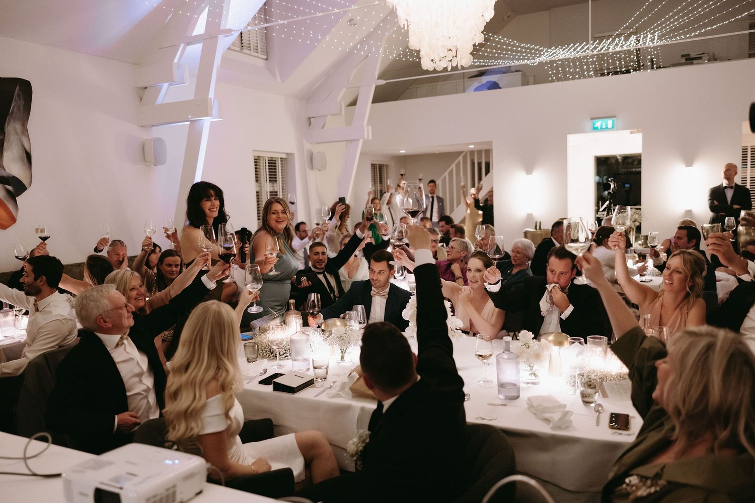 A series of candlelit dinner tables filled with guests at an elegant white event space with high ceilings.
