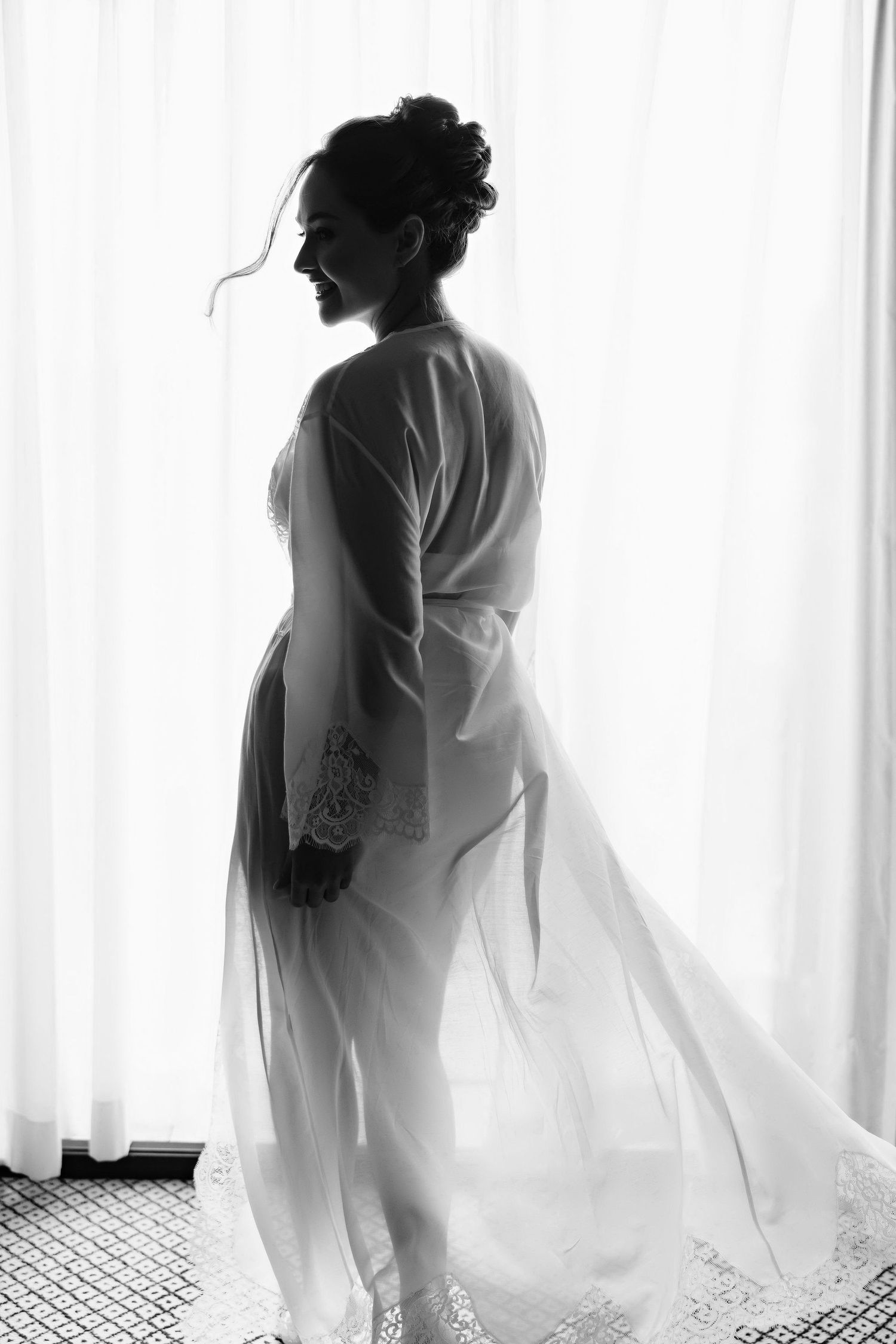 Black and white silhouette of a pregnant person in a flowing white robe standing by a window with sheer curtains.