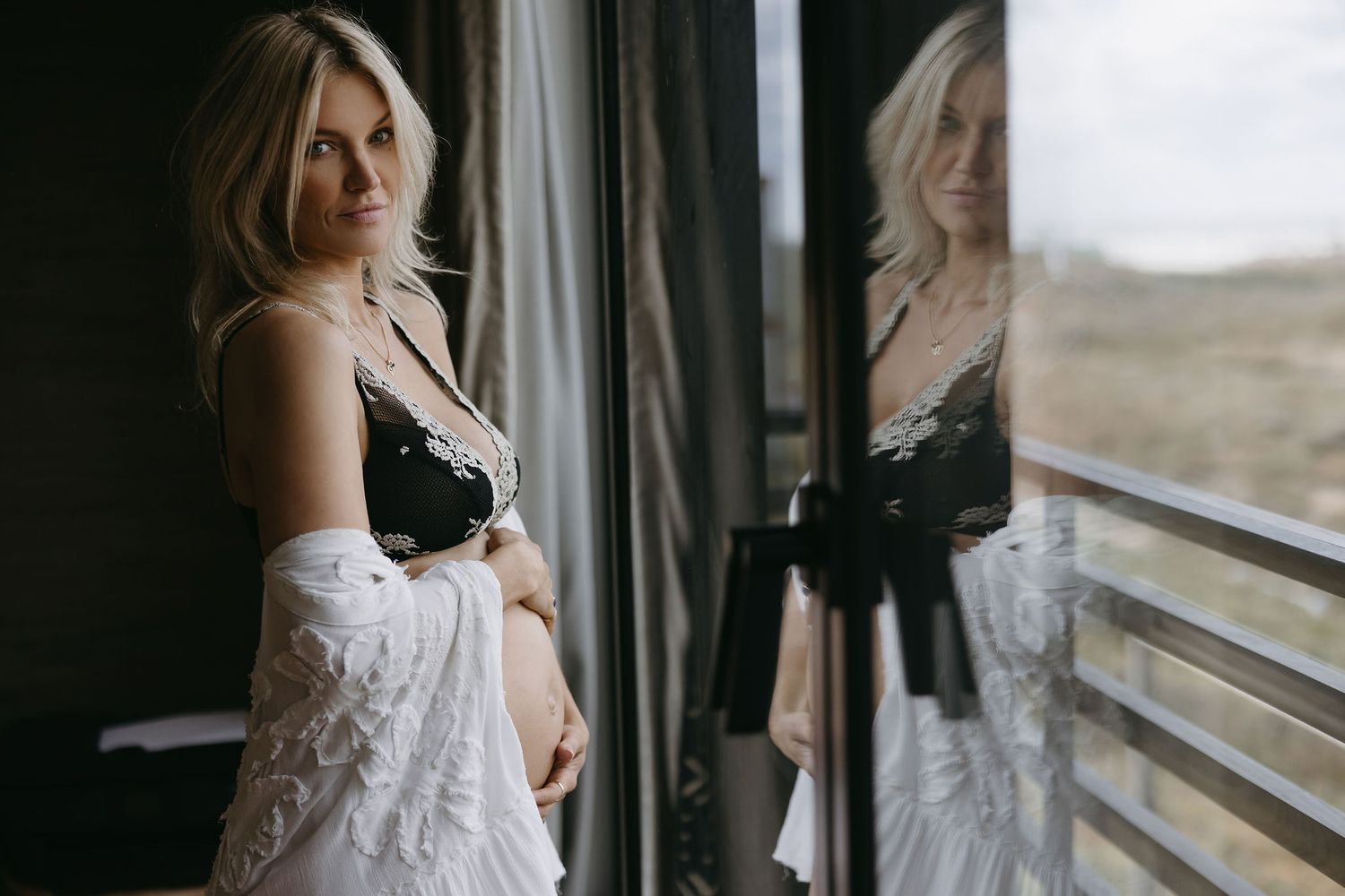 Woman in white lace dress looking out window during intimate photoshoot.