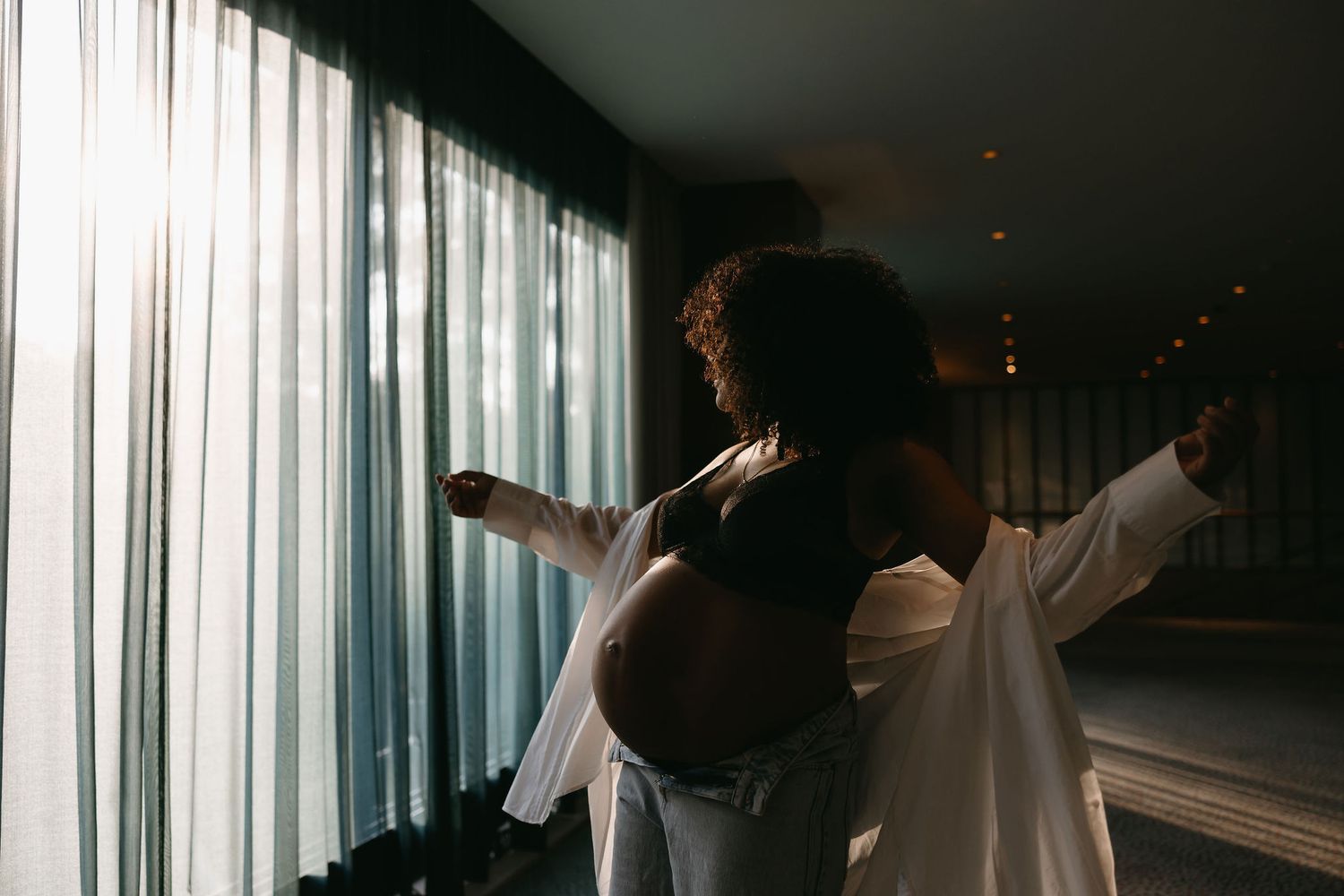 Silhouette of pregnant figure by window in flowing white clothing.