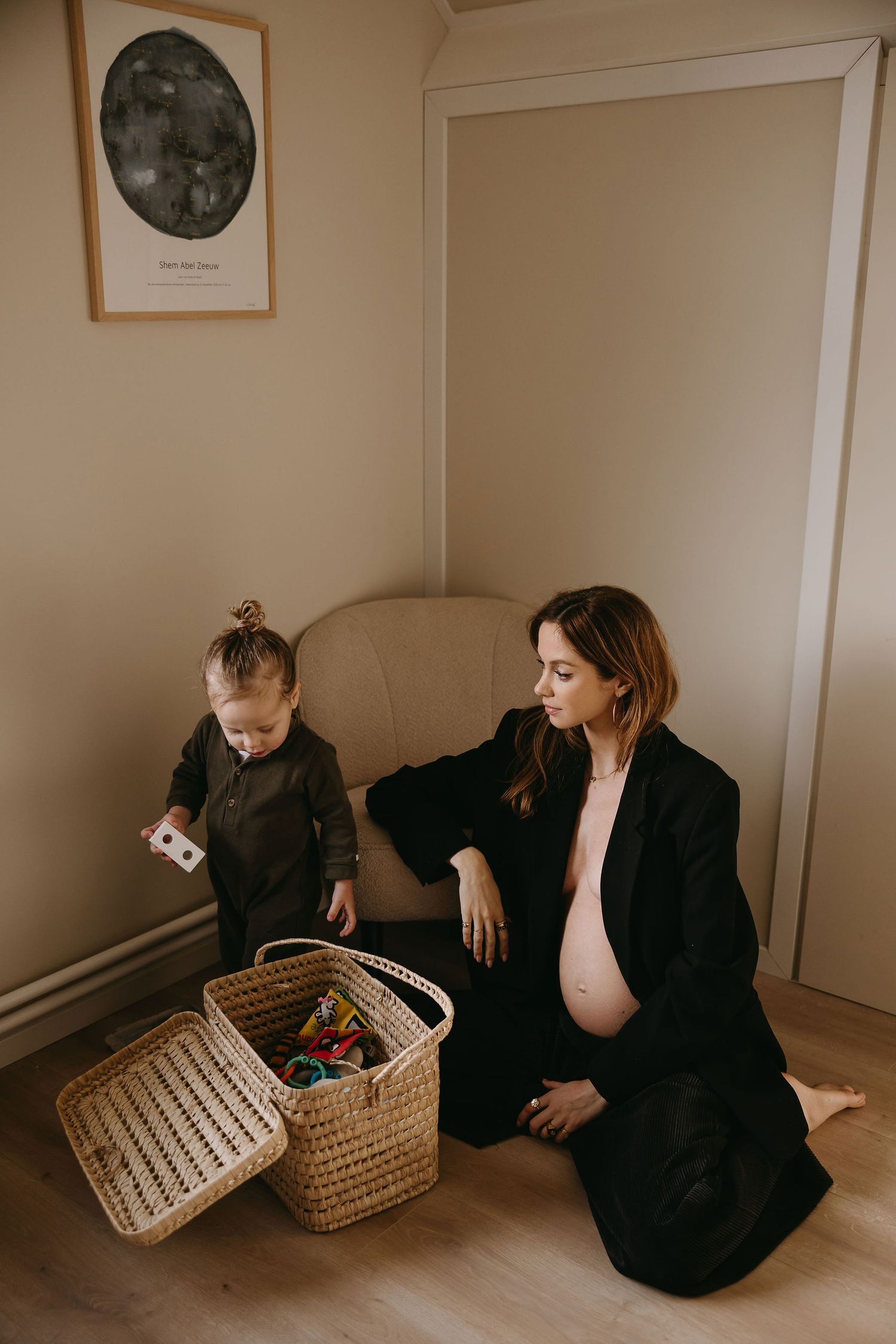 A pregnant woman in black coat sits beside a wicker basket while a child explores.