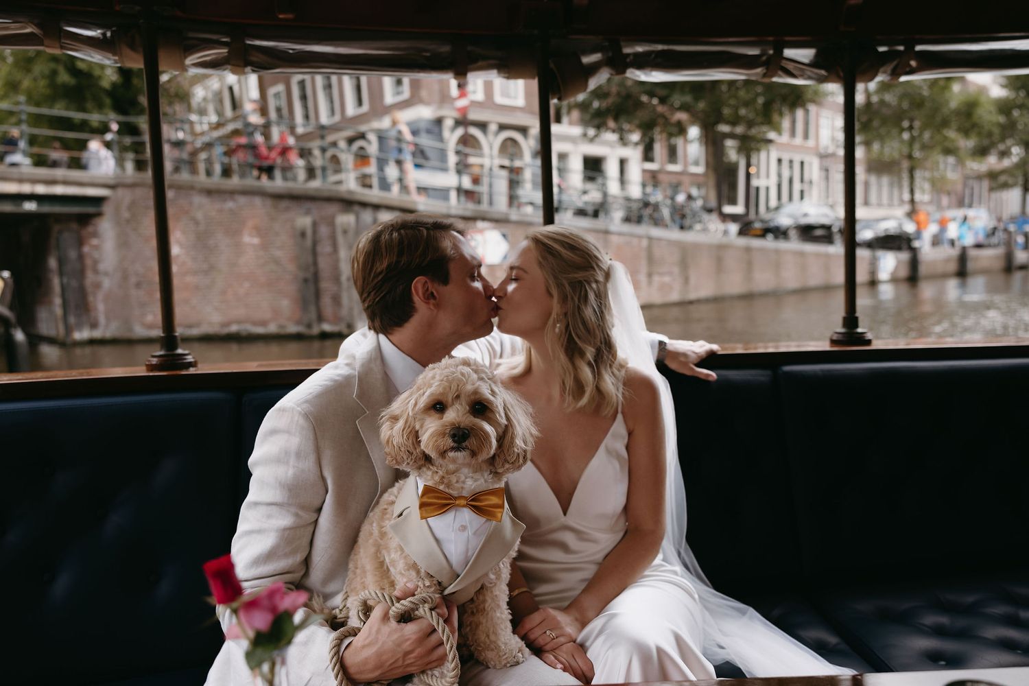 A small dog wearing a bowtie sits in a boat with Amsterdam canals in the background.