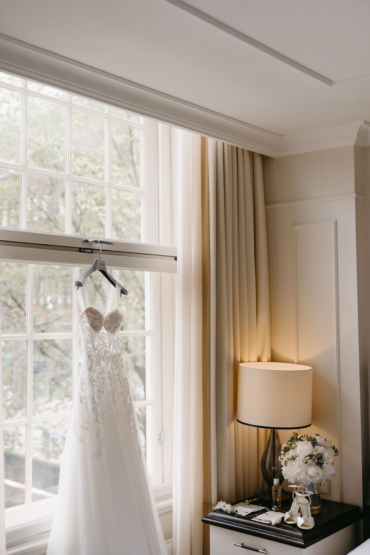 White lace wedding dress hanging by a window with a table lamp and floral bouquet on side table.