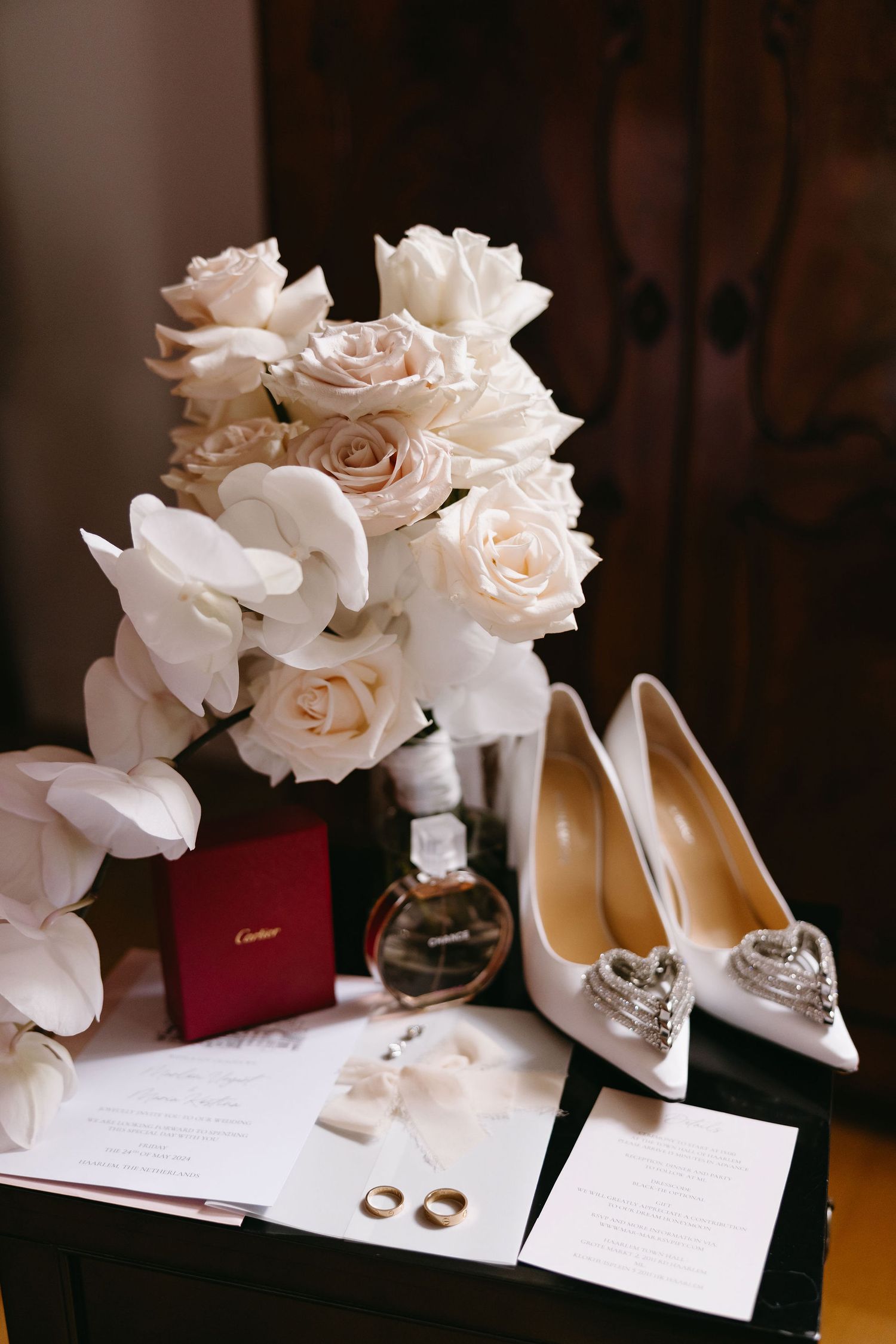 Wedding details flatlay with white roses, elegant heels, rings and invitation on light surface.