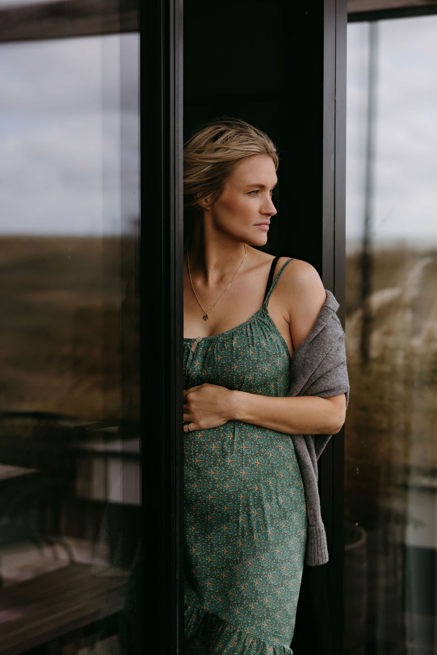 Person in a green floral dress gazing through window with scenic countryside view.