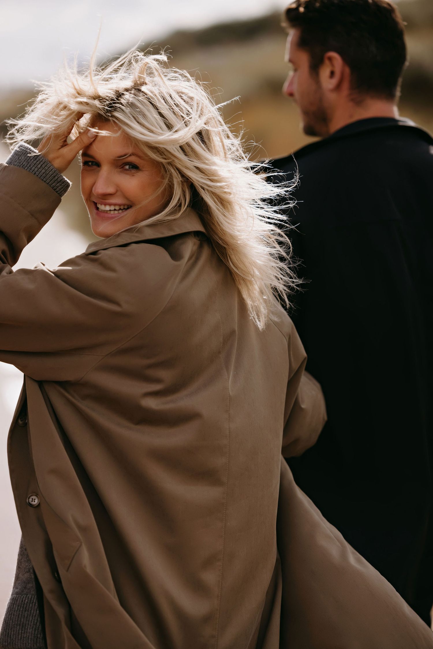 A woman in a tan coat turns to smile as wind blows through her hair on a cloudy day.