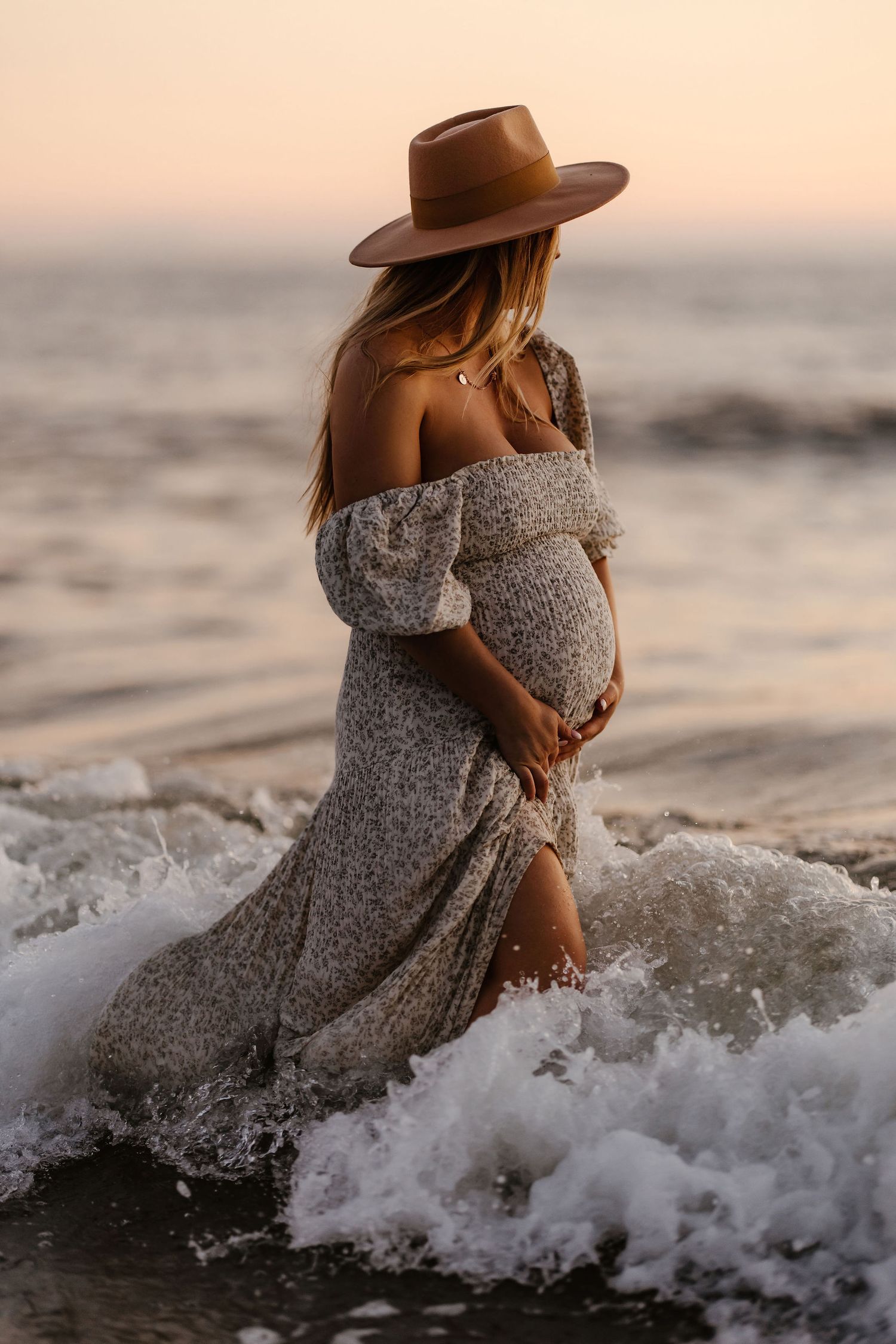 Silhouette of pregnant woman in flowing dress and hat standing in ocean waves at sunset.
