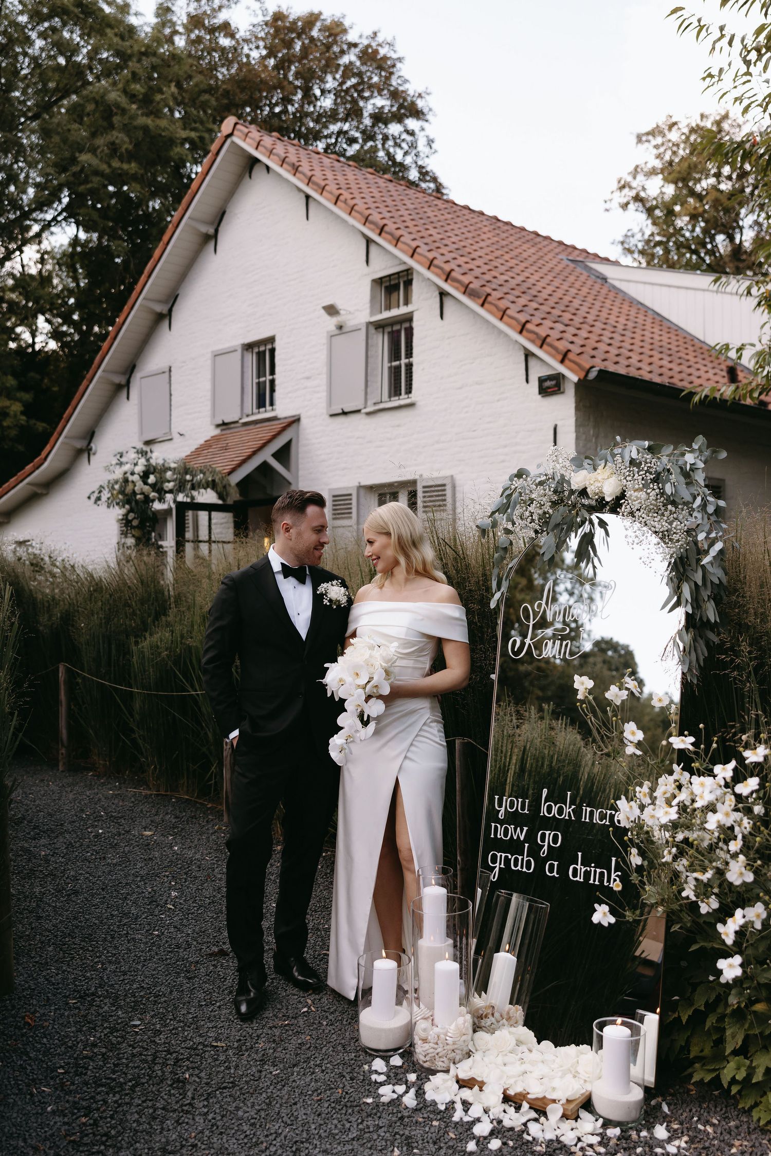 Wedding couple stands outside white house with terra cotta roof, surrounded by romantic candlelight decor.