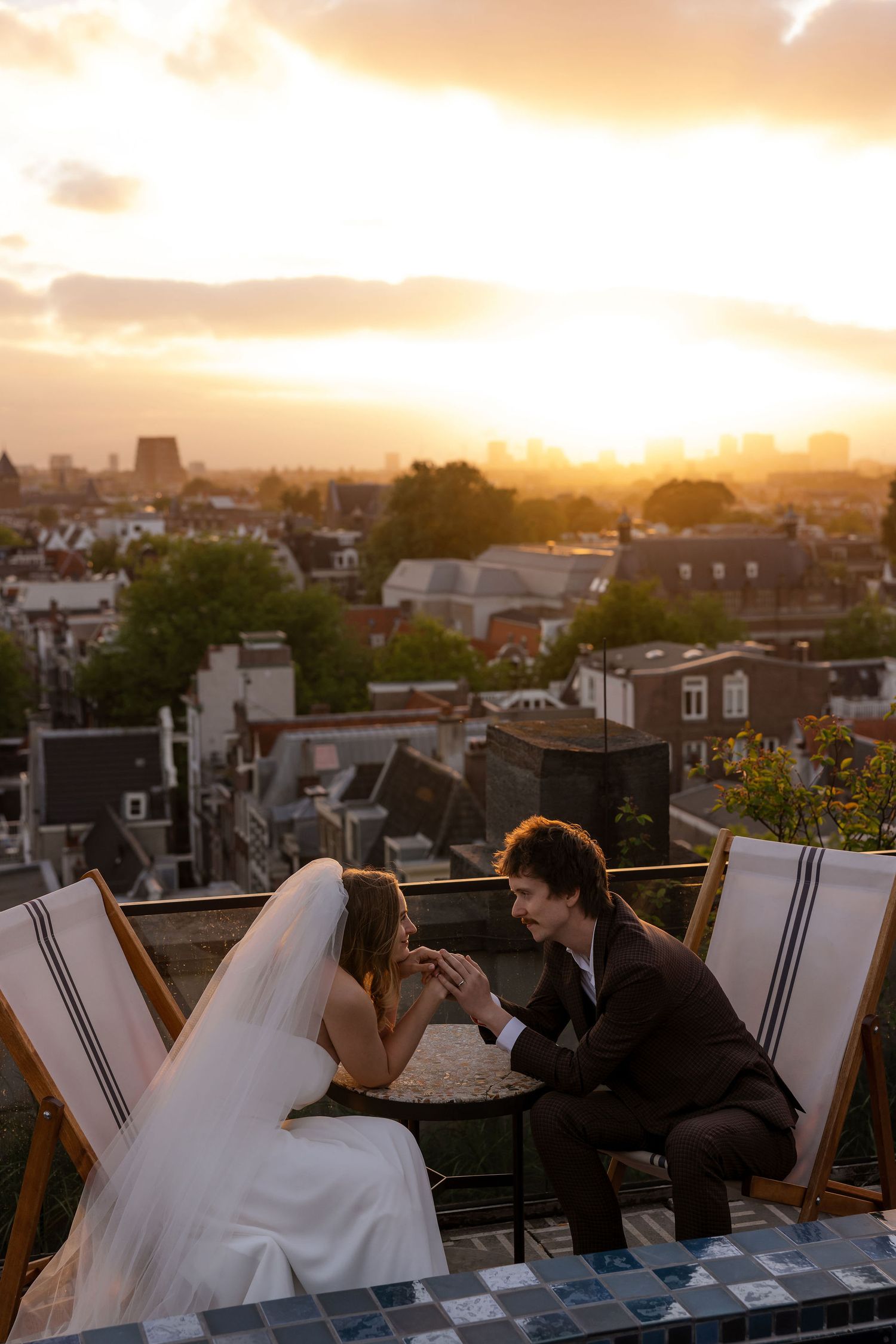 A wedding couple and guests paint on easels while watching a golden sunset over a rooftop cityscape.