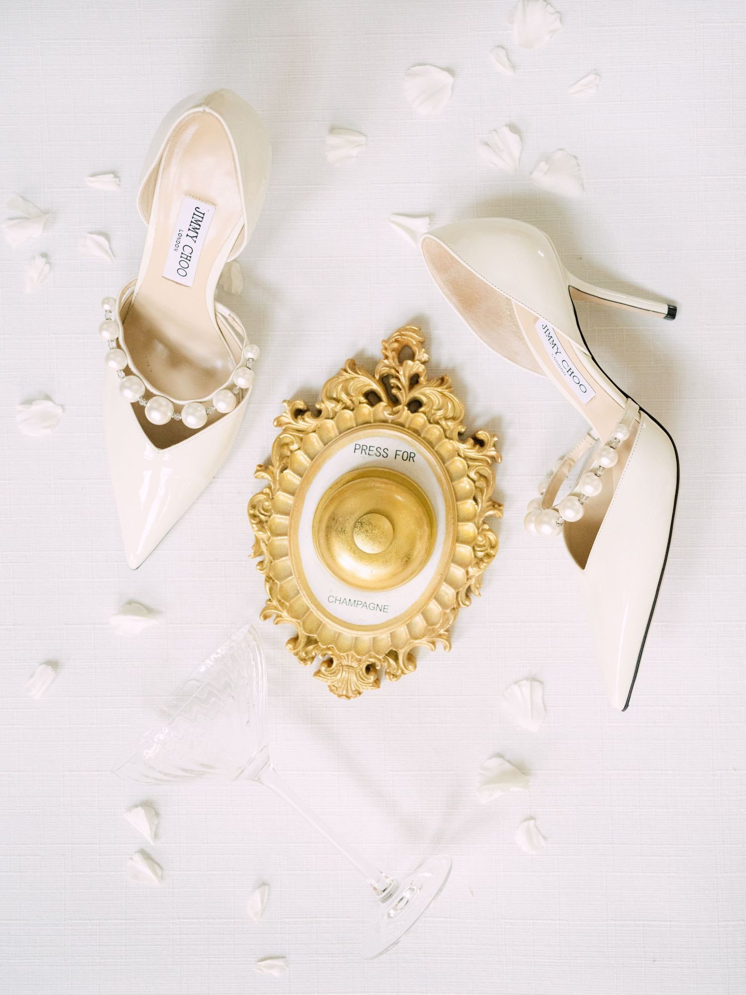 Wedding flatlay featuring Jimmy Choo bridal shoes paired with a chic 'Press for Champagne' bell, styled for a luxury wedding detail shot