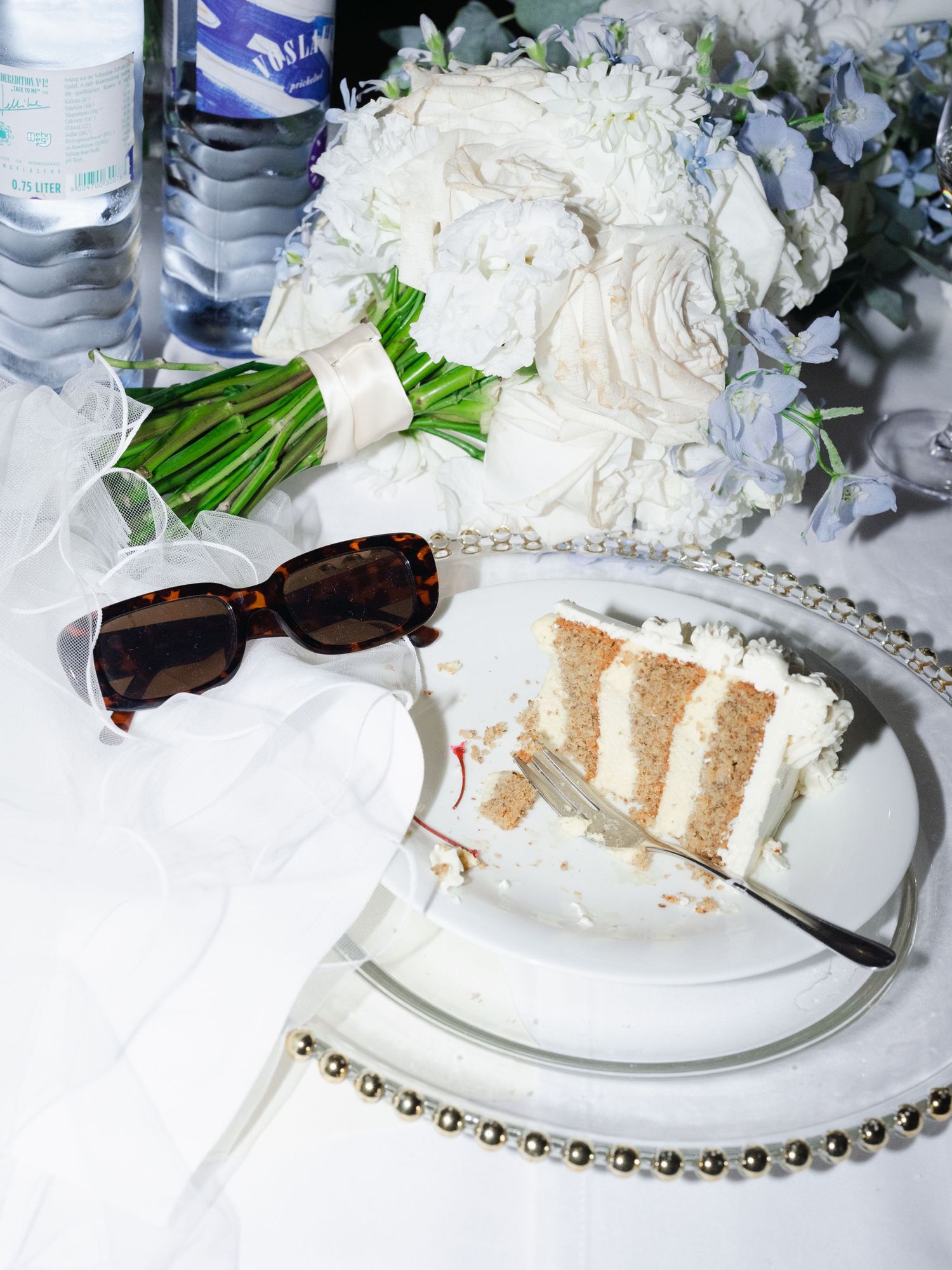 Wedding party flatlay with chic sunglasses, vibrant bridal bouquet, and partially eaten cake, styled for a modern luxury wedding detail shot
