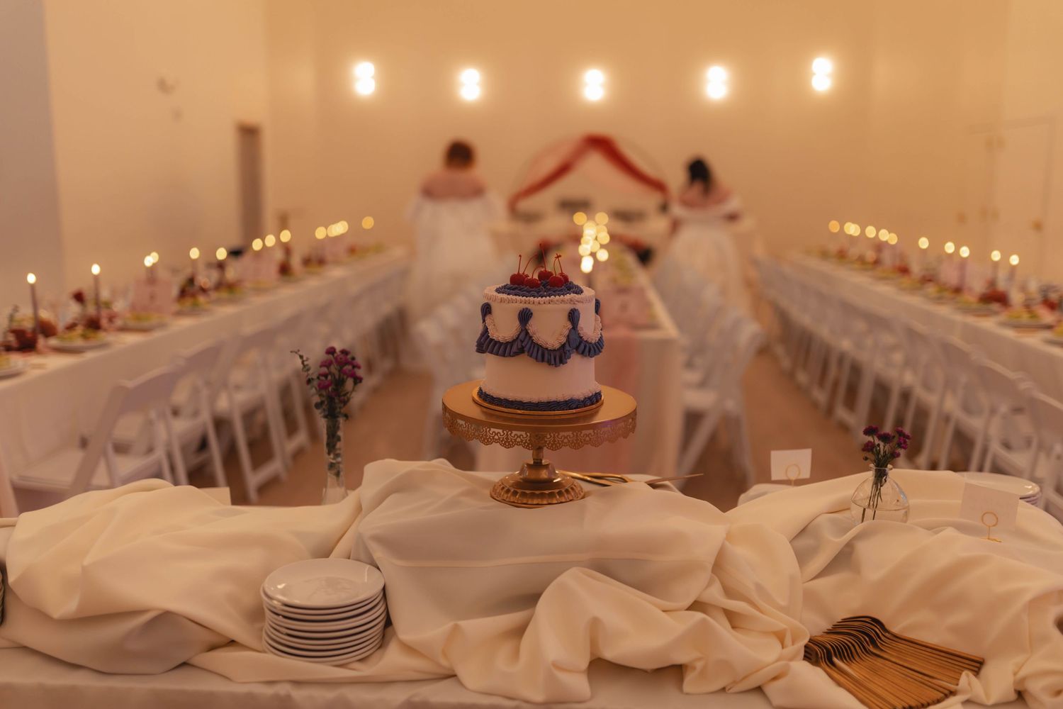 Elegant wedding reception hall with a decorated blue and white cake on a gold stand, long tables with candles and white linens.