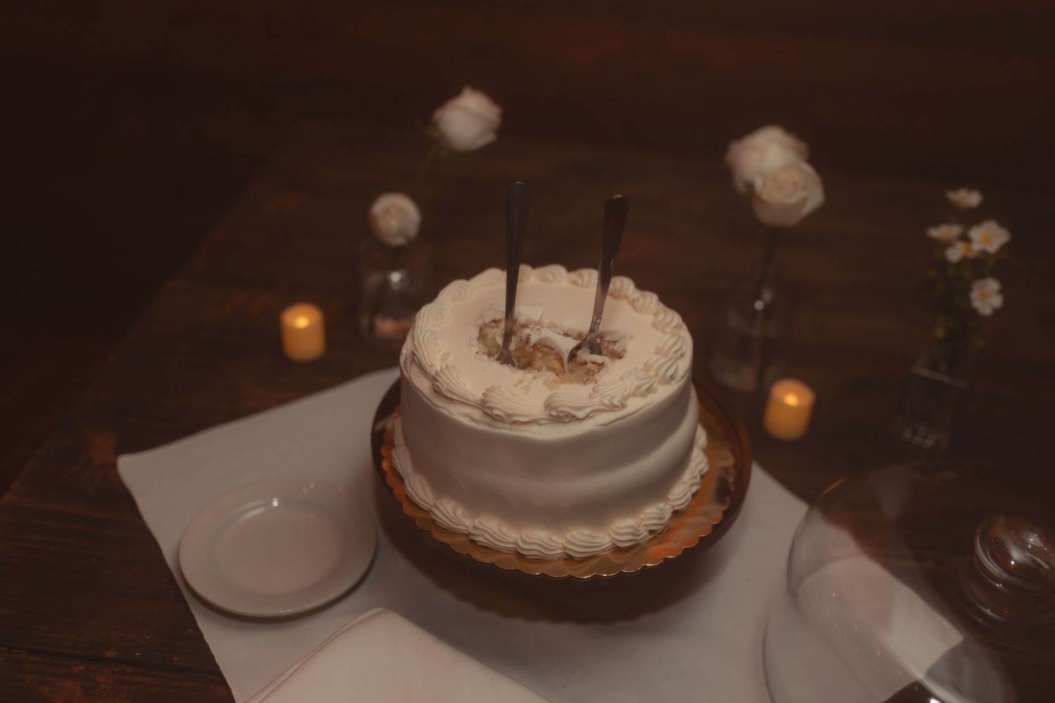 Elegant two-tier white wedding cake with candles on a wooden table surrounded by glowing candles and floral accents.