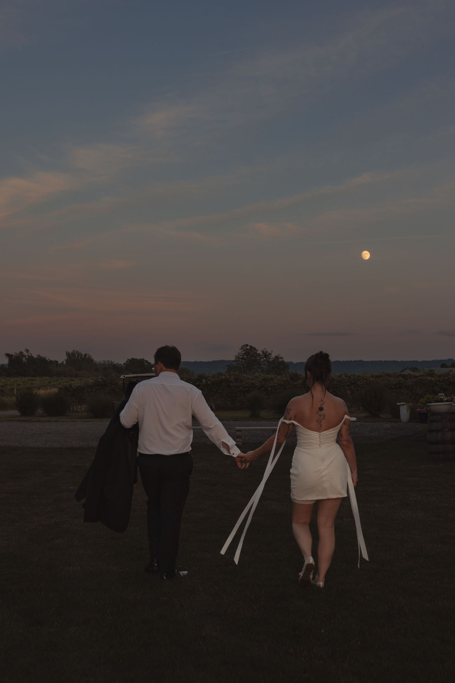 Couple in wedding attire holding hands walking toward full moon at dusk on grassy field.
