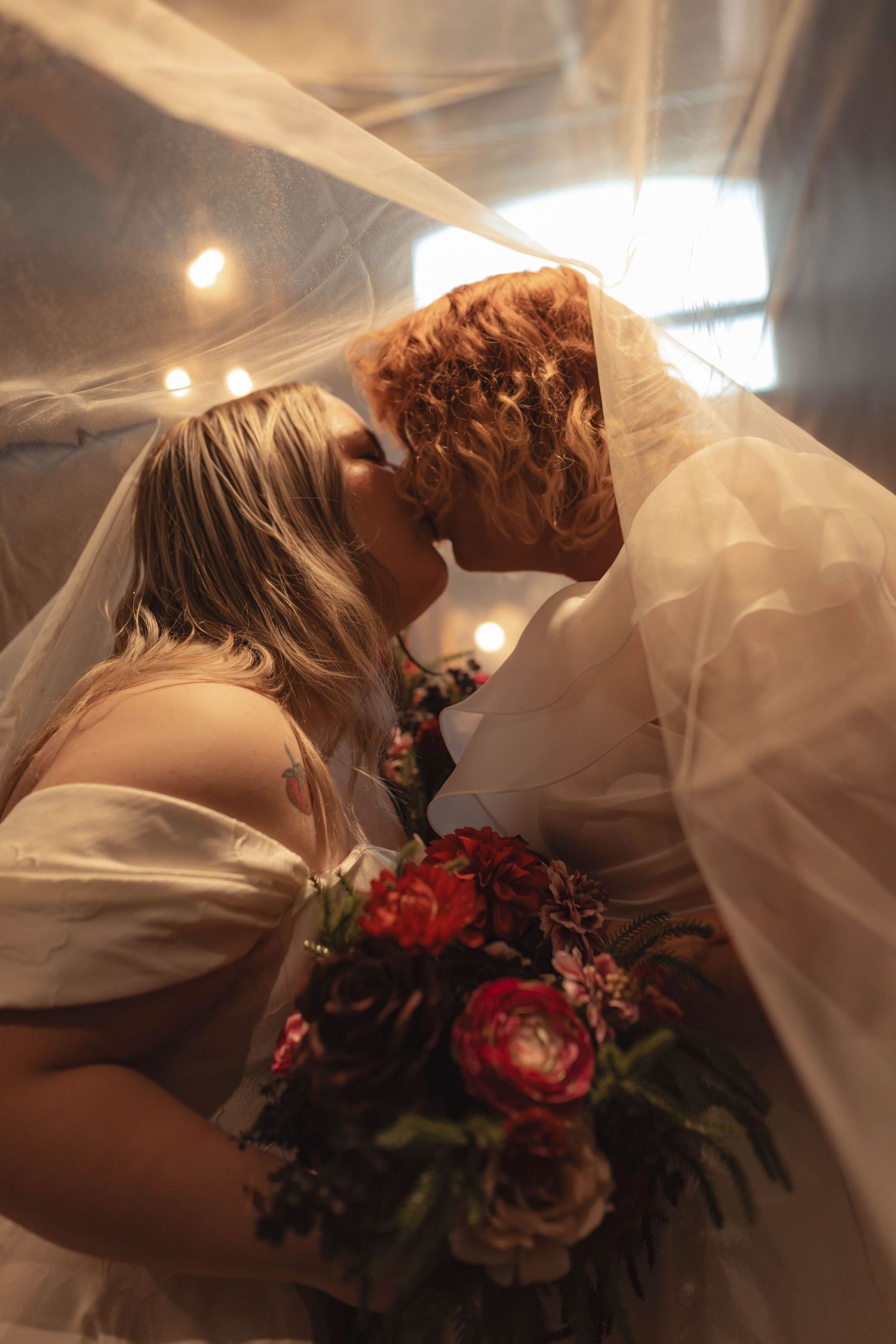 Two brides sharing a romantic kiss under a veil with soft bokeh lights and a red bridal bouquet.