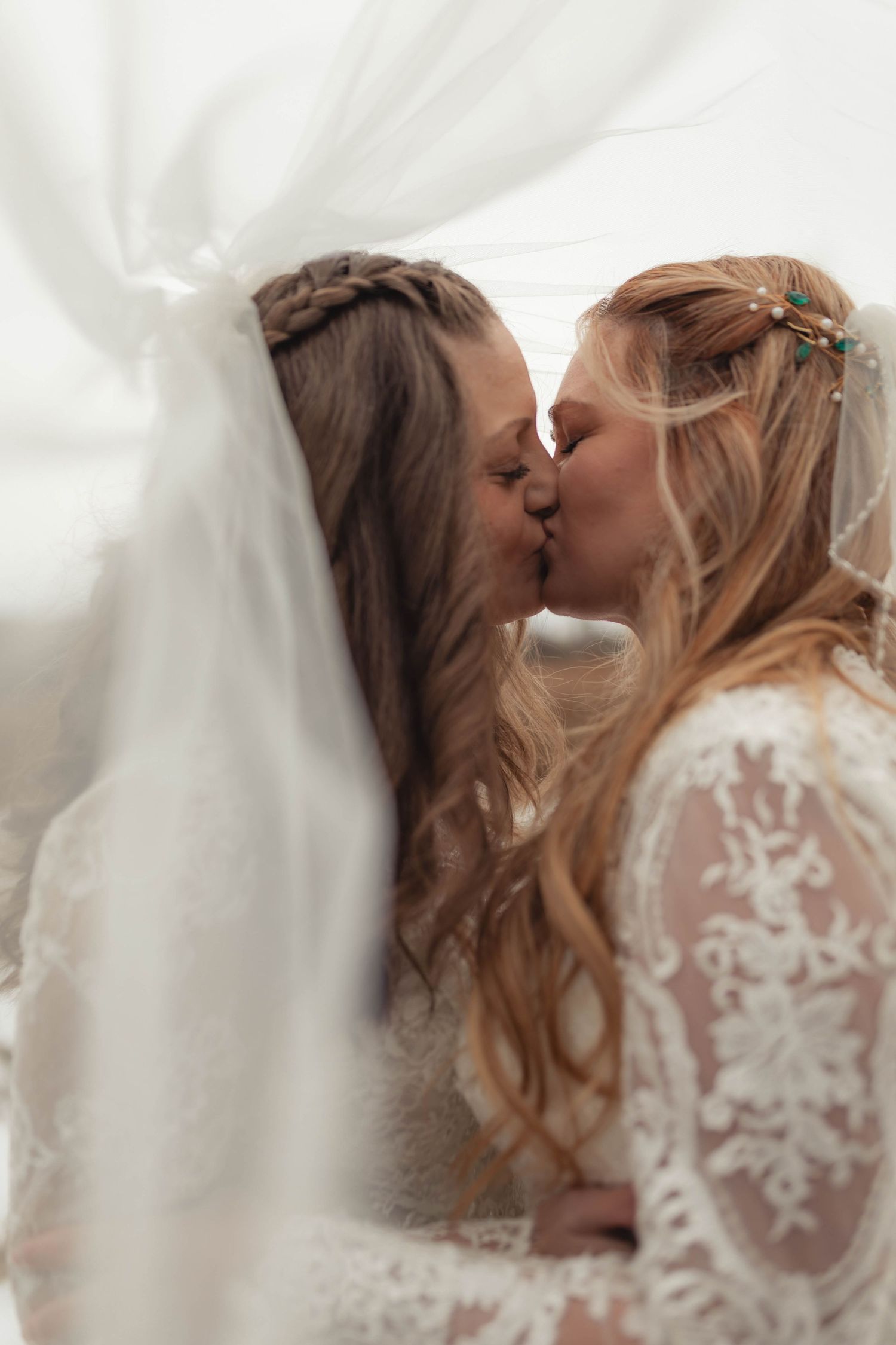 Two brides sharing a romantic kiss beneath a flowing white veil on their wedding day.