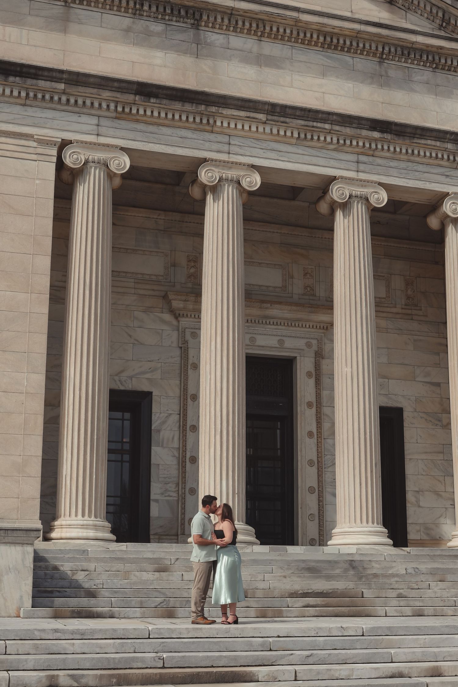 Couple embracing on steps of neoclassical building with tall Ionic columns in warm sunlight.