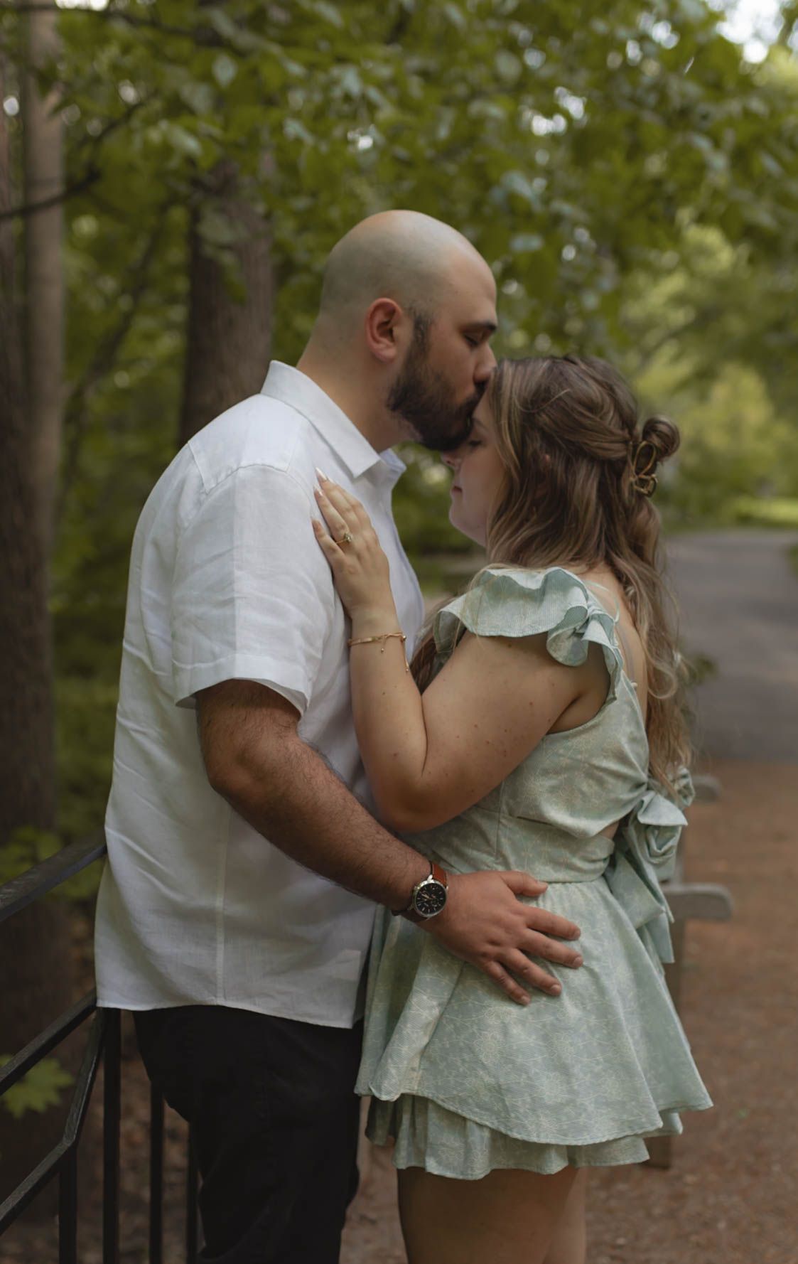 Couple sharing a kiss on a tree-lined park path, woman wearing a mint ruffle dress.