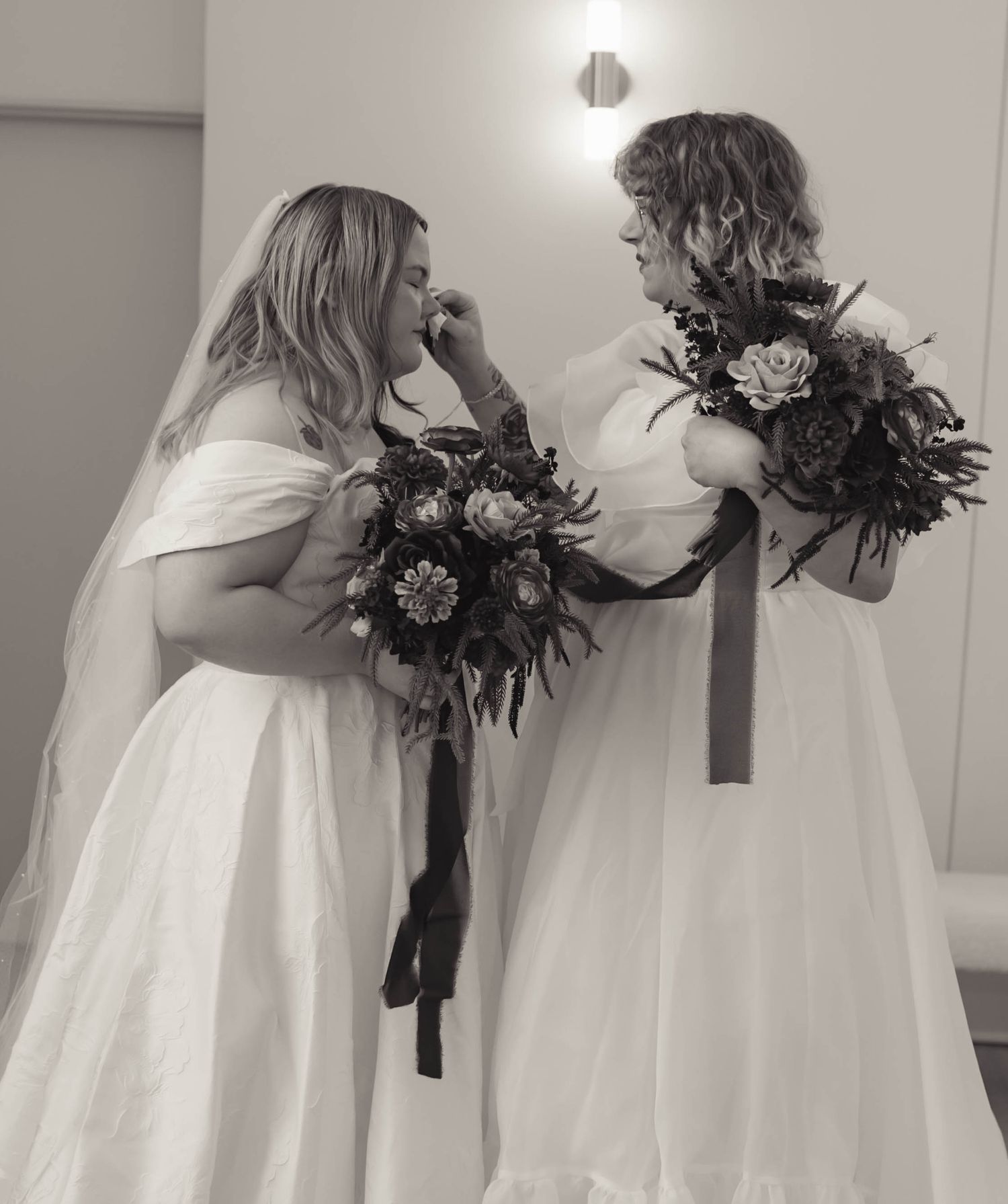 Two brides in white wedding gowns share an emotional moment holding floral bouquets in black and white photo.