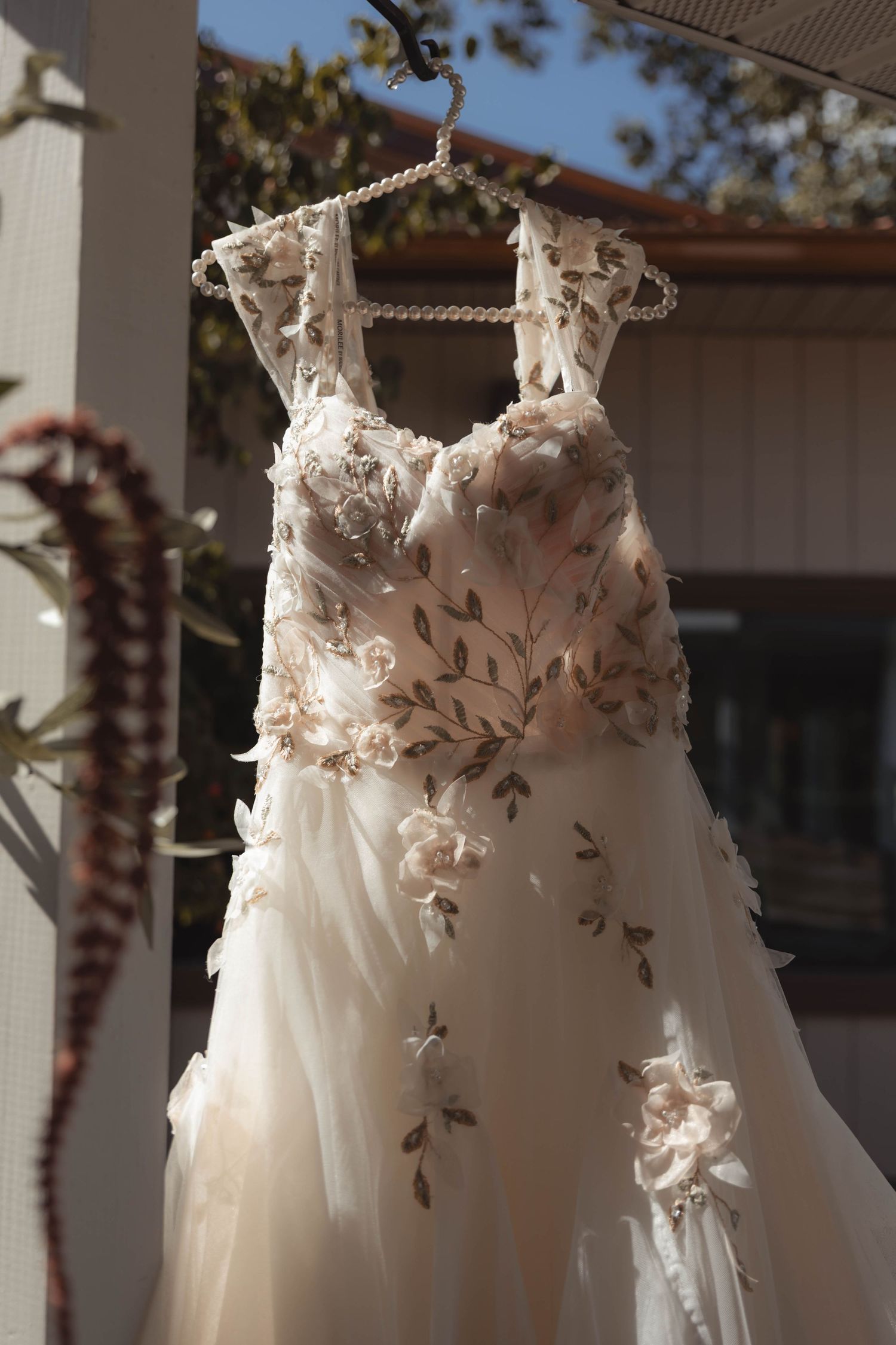 Elegant floral embroidered wedding dress with tulle skirt hanging on a pearl hanger outdoors in sunlight.
