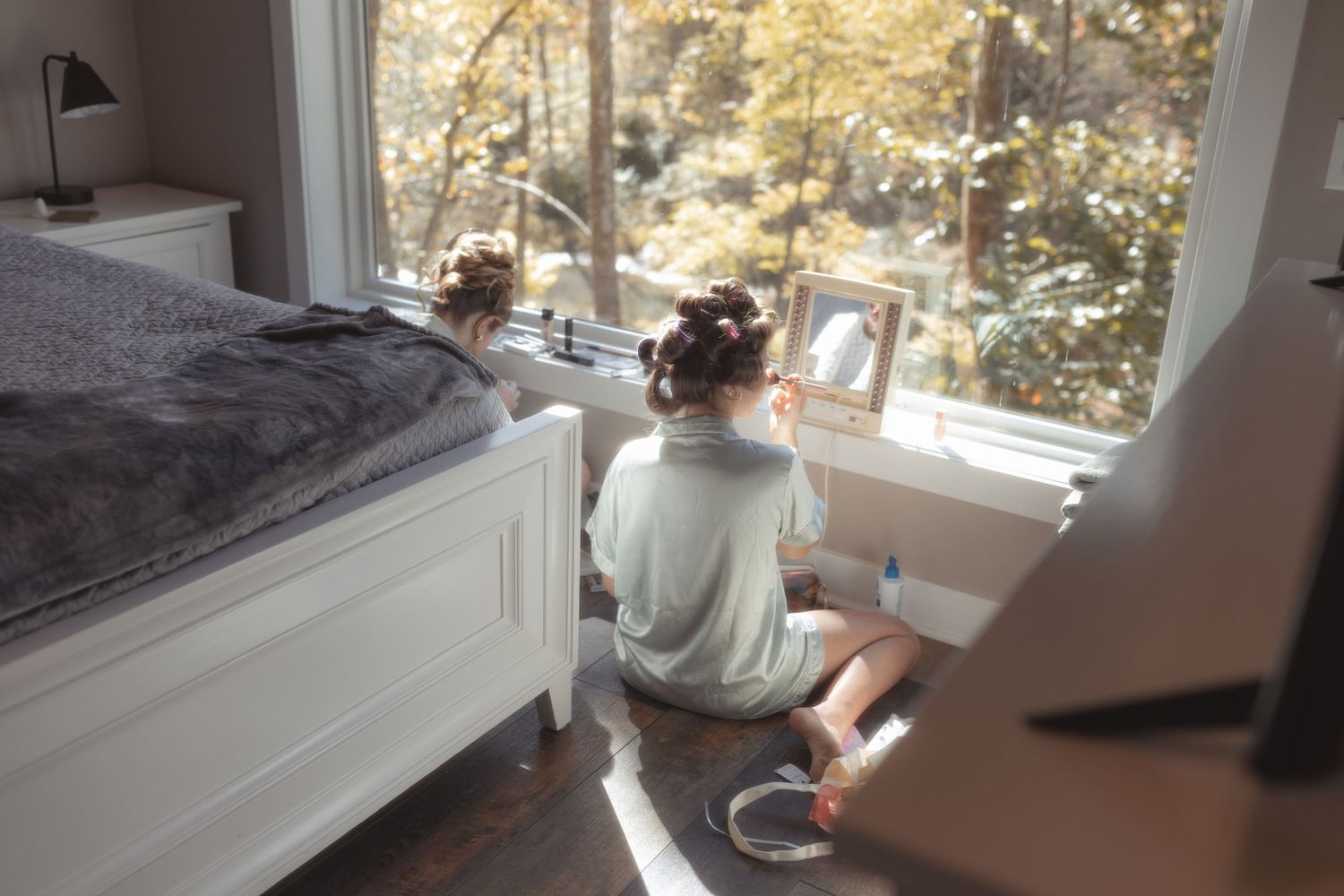 Bride sitting on bedroom floor in morning light, preparing for wedding day while looking out large window.