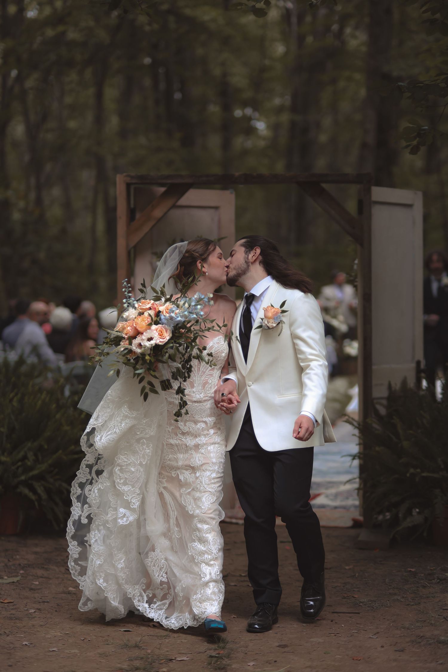 Bride and groom share first kiss at outdoor forest wedding, bride holding peach floral bouquet in lace gown.