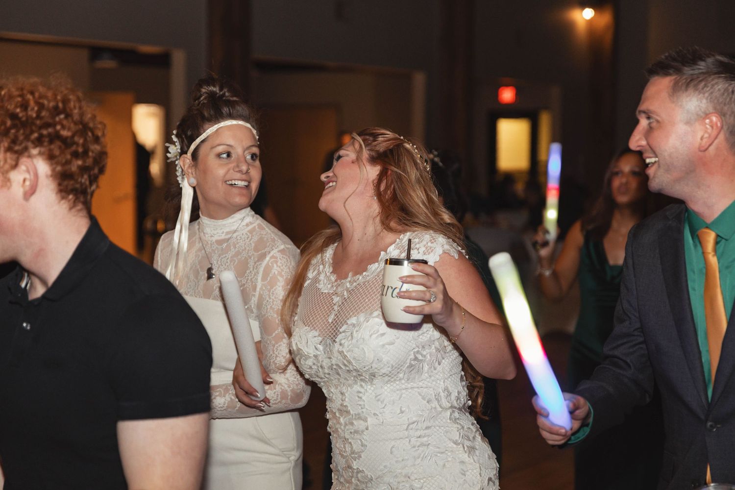 Two brides laughing and celebrating with guests holding glowsticks at wedding reception.