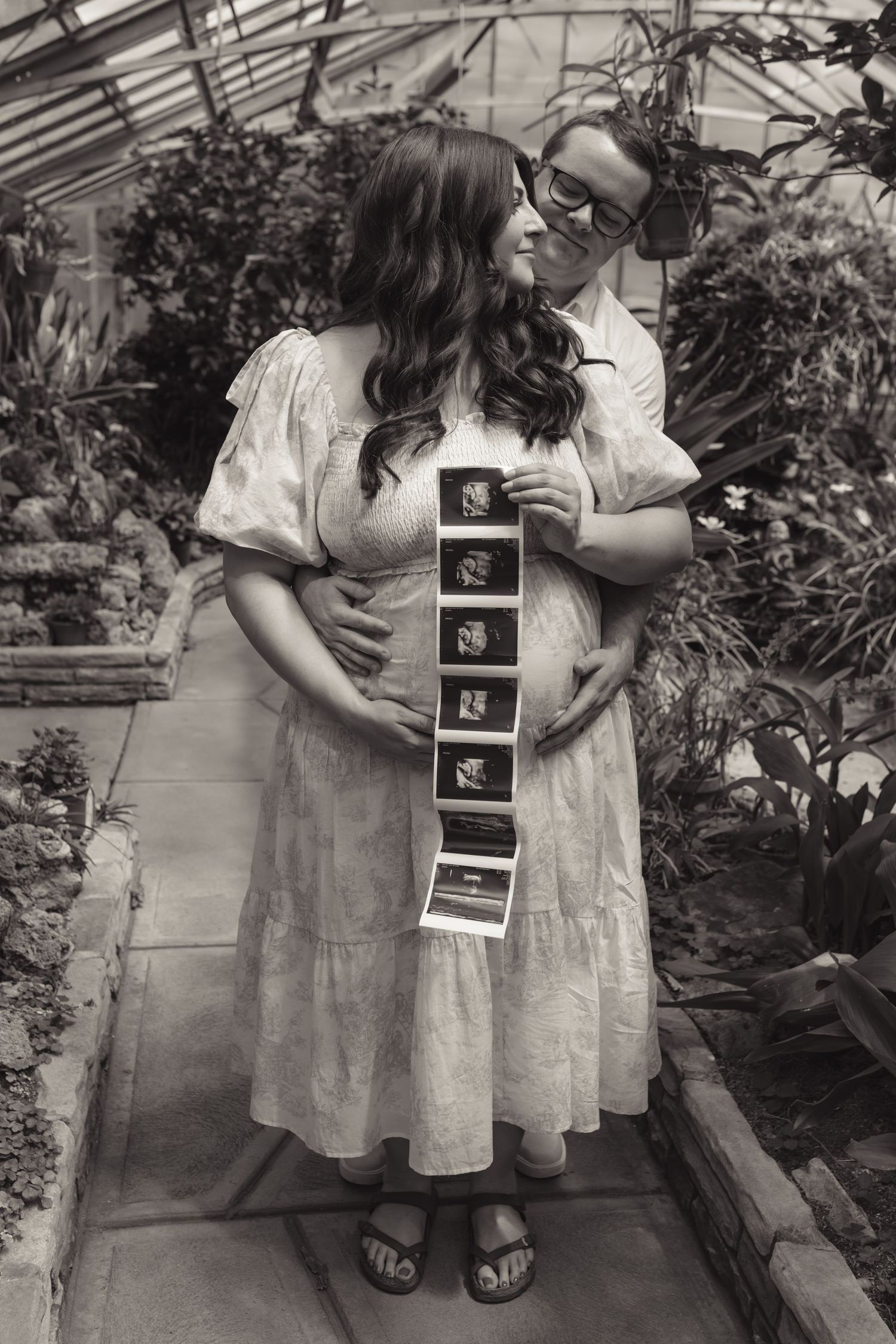 Black and white photo of pregnant couple holding ultrasound strip in a lush greenhouse, celebrating their upcoming arrival.