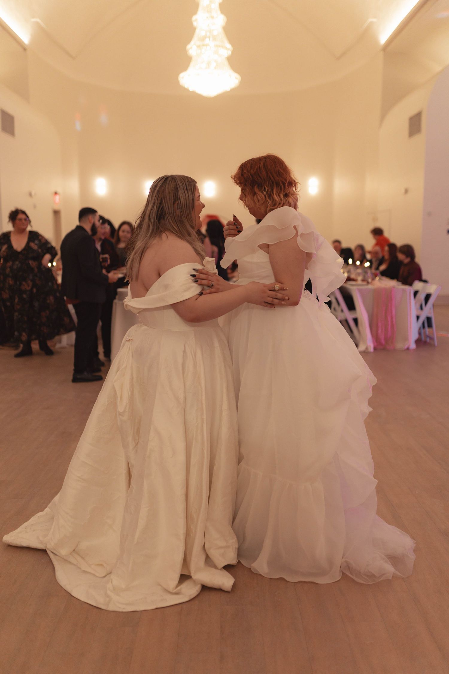 Two brides in white gowns share a dance at their wedding reception in an elegant ballroom.