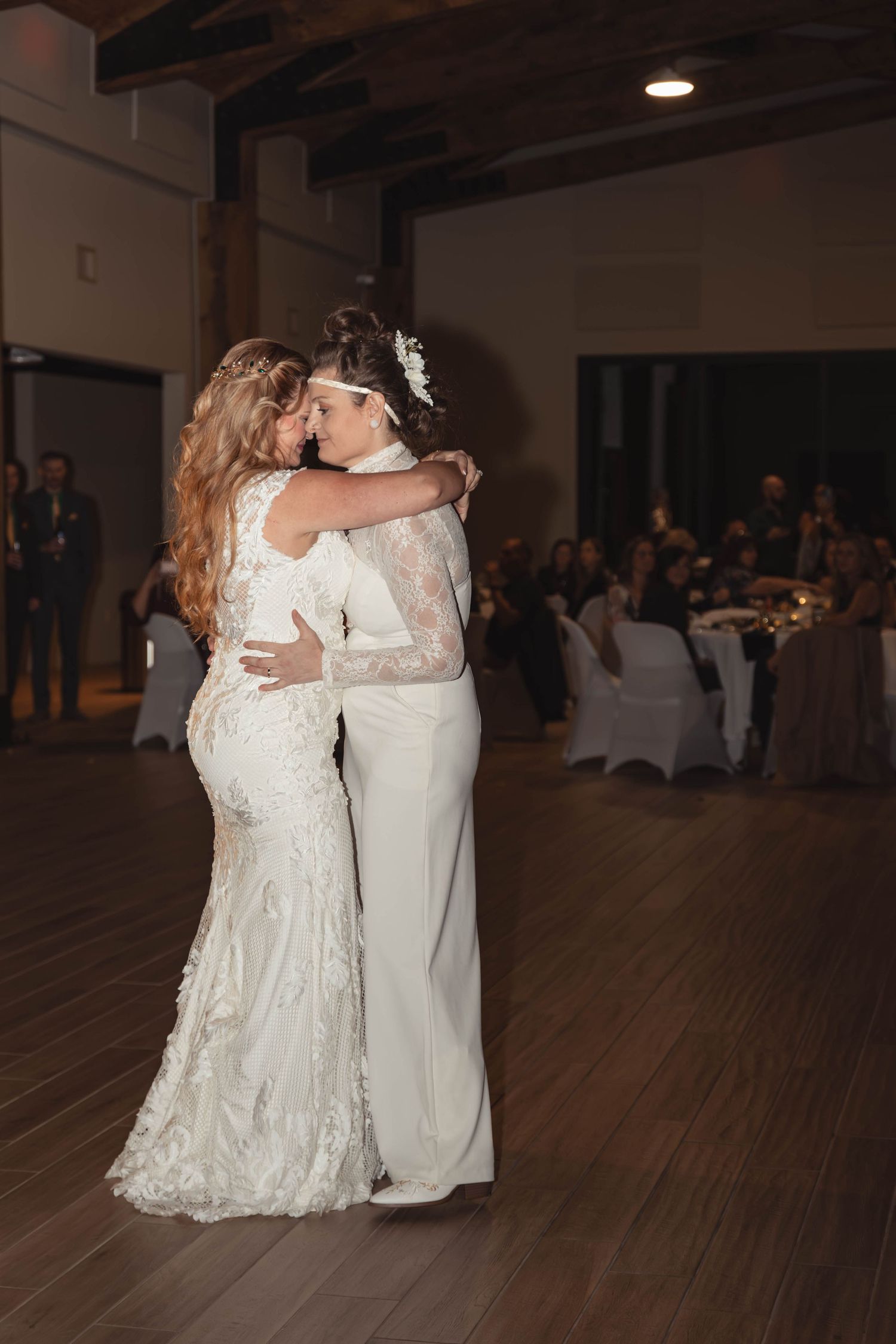 Two brides sharing their first dance in elegant white wedding attire at a romantic indoor reception venue.