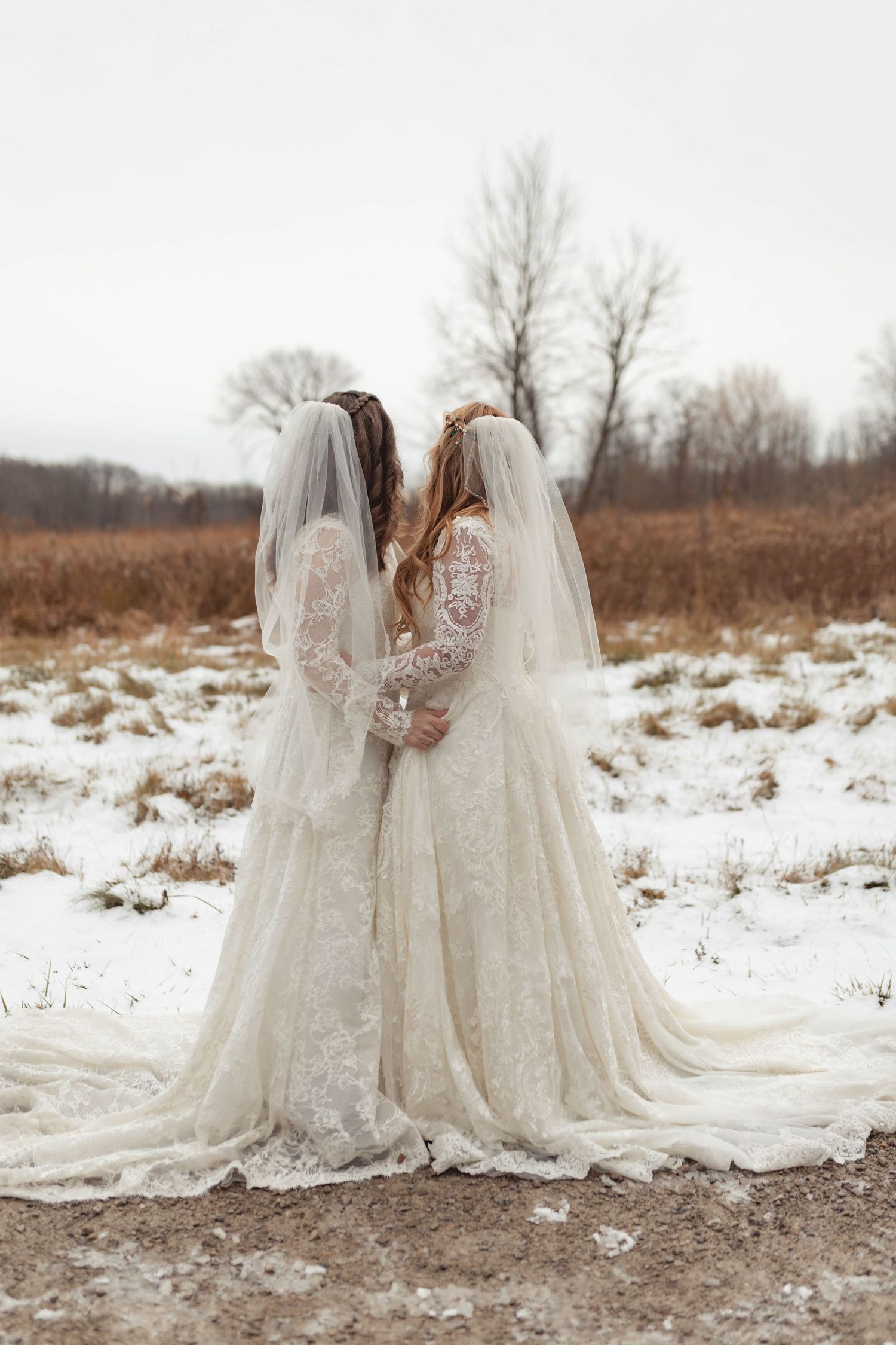 Two brides in white lace wedding gowns with veils stand together in a snowy winter field.