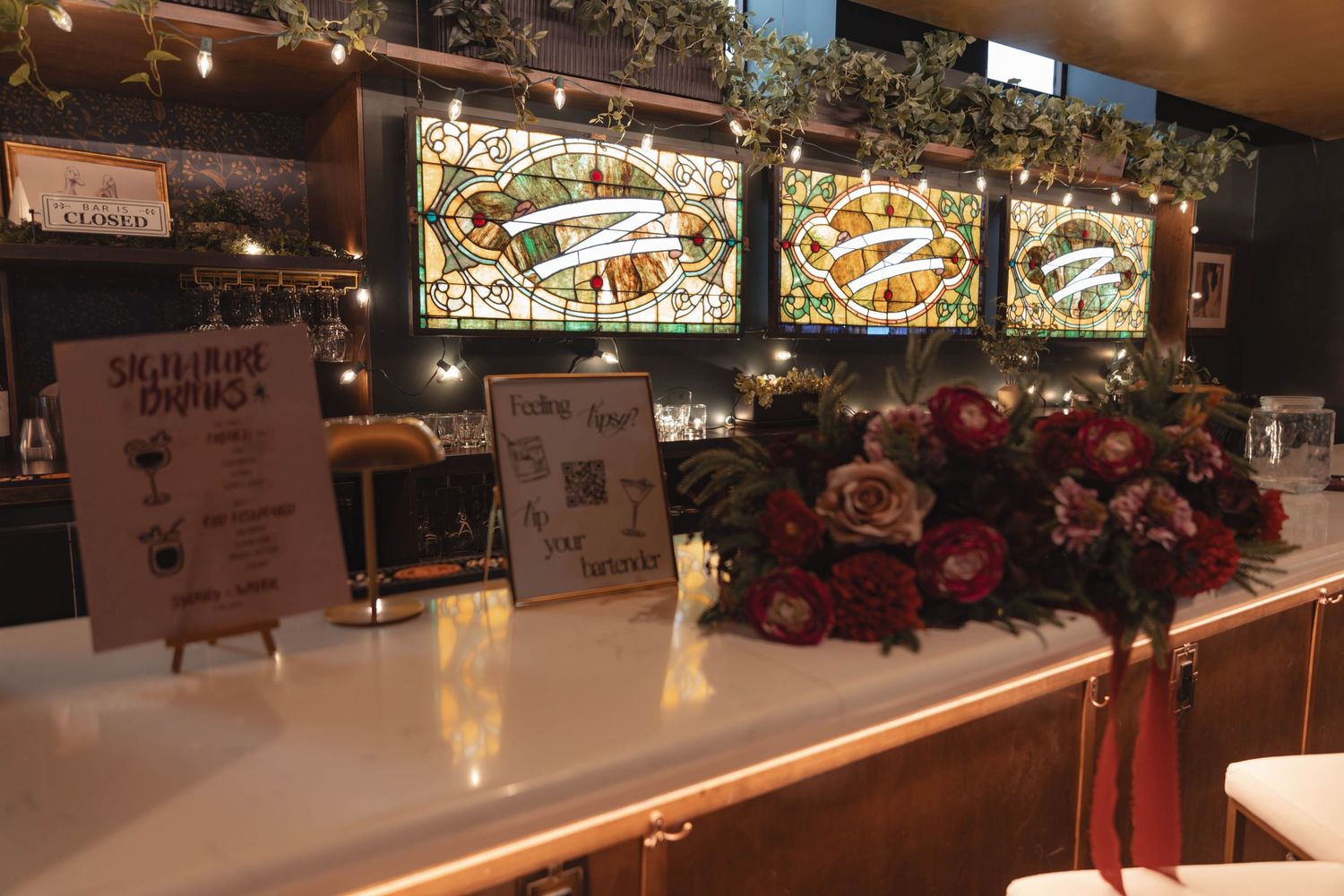 Wedding reception bar decorated with red and white floral arrangement and stained glass backdrop.