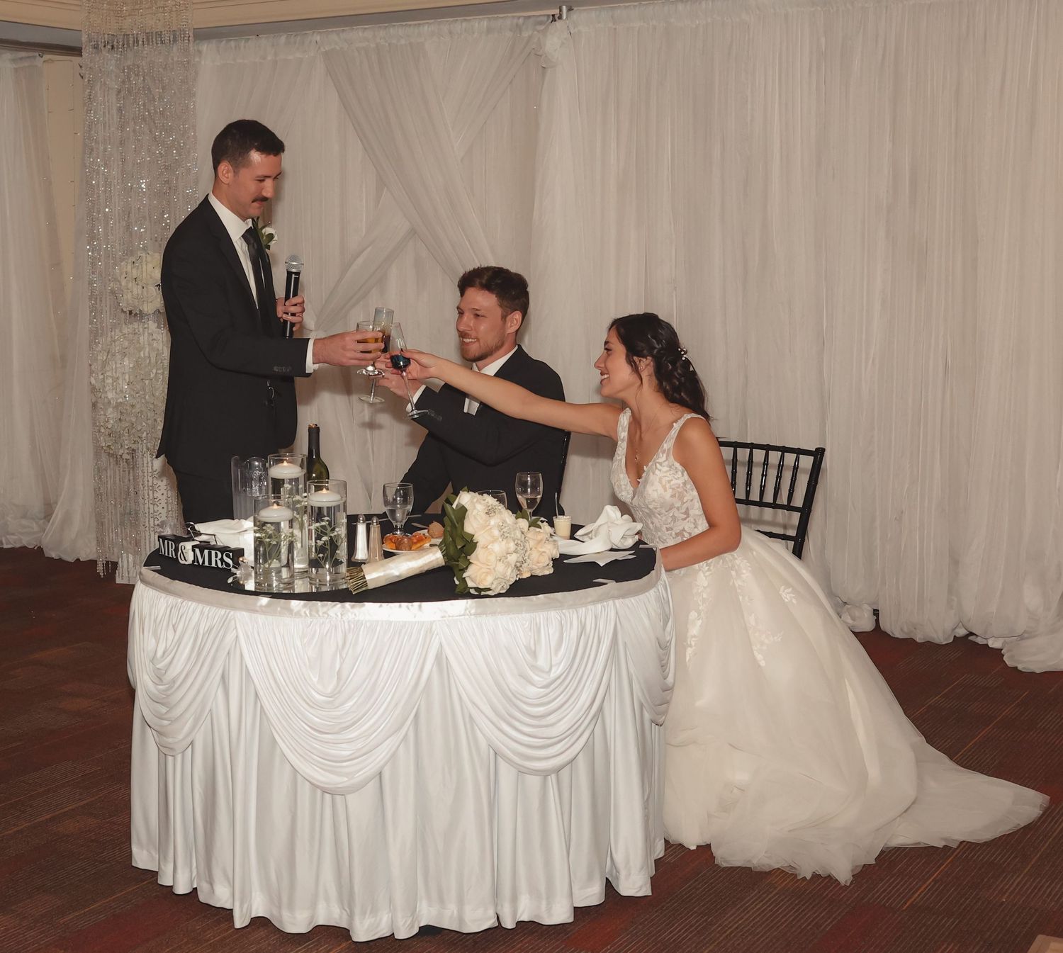 Best man gives a toast as bride and groom raise glasses at elegantly decorated sweetheart table at reception.