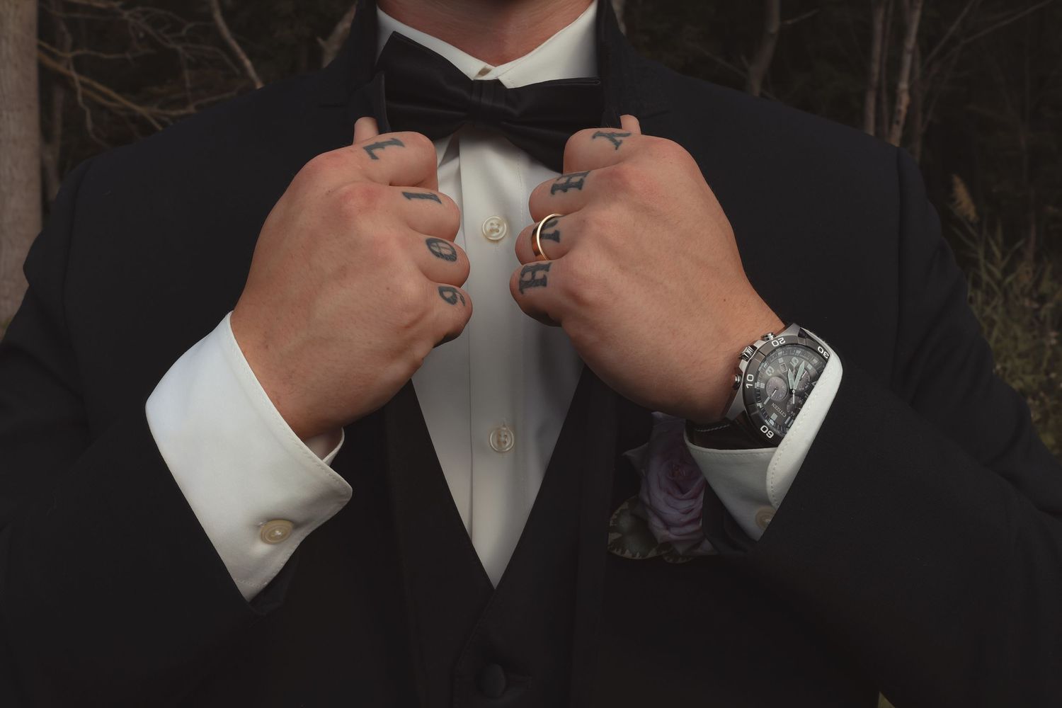 Man in black tuxedo with bow tie adjusting lapels, wearing rings and a decorative wristwatch.