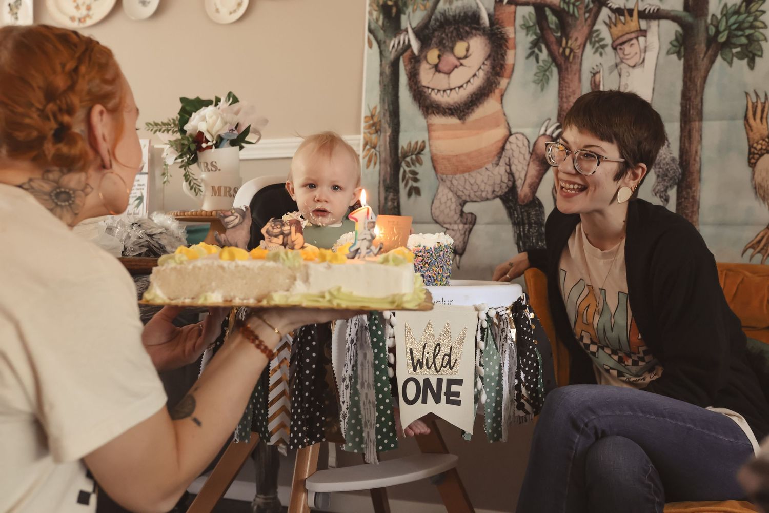Baby in a Wild One high chair celebrates first birthday with two women presenting a decorated cake.