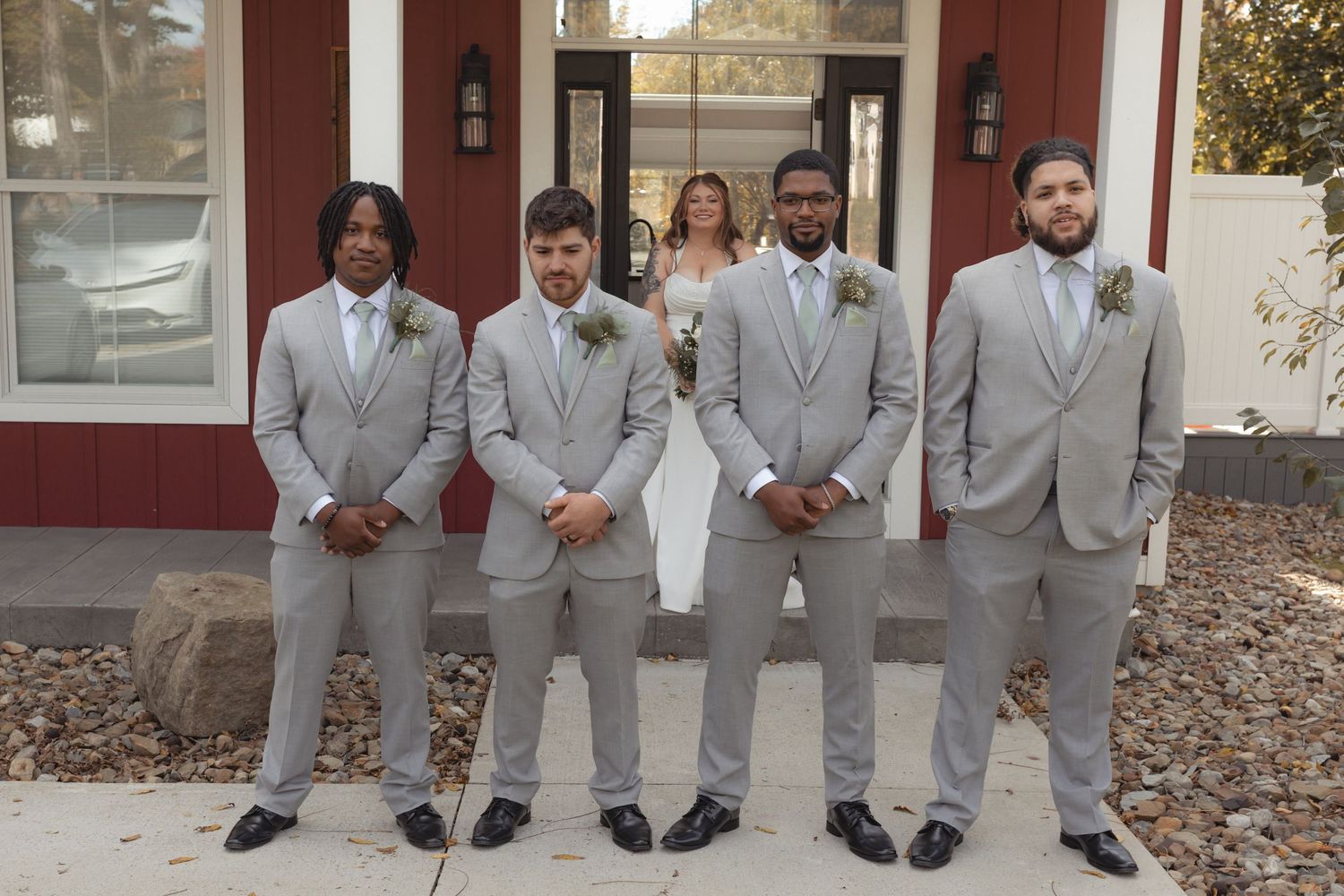Four groomsmen in gray suits with boutonnieres stand outside a red building, with a bride visible in the background.