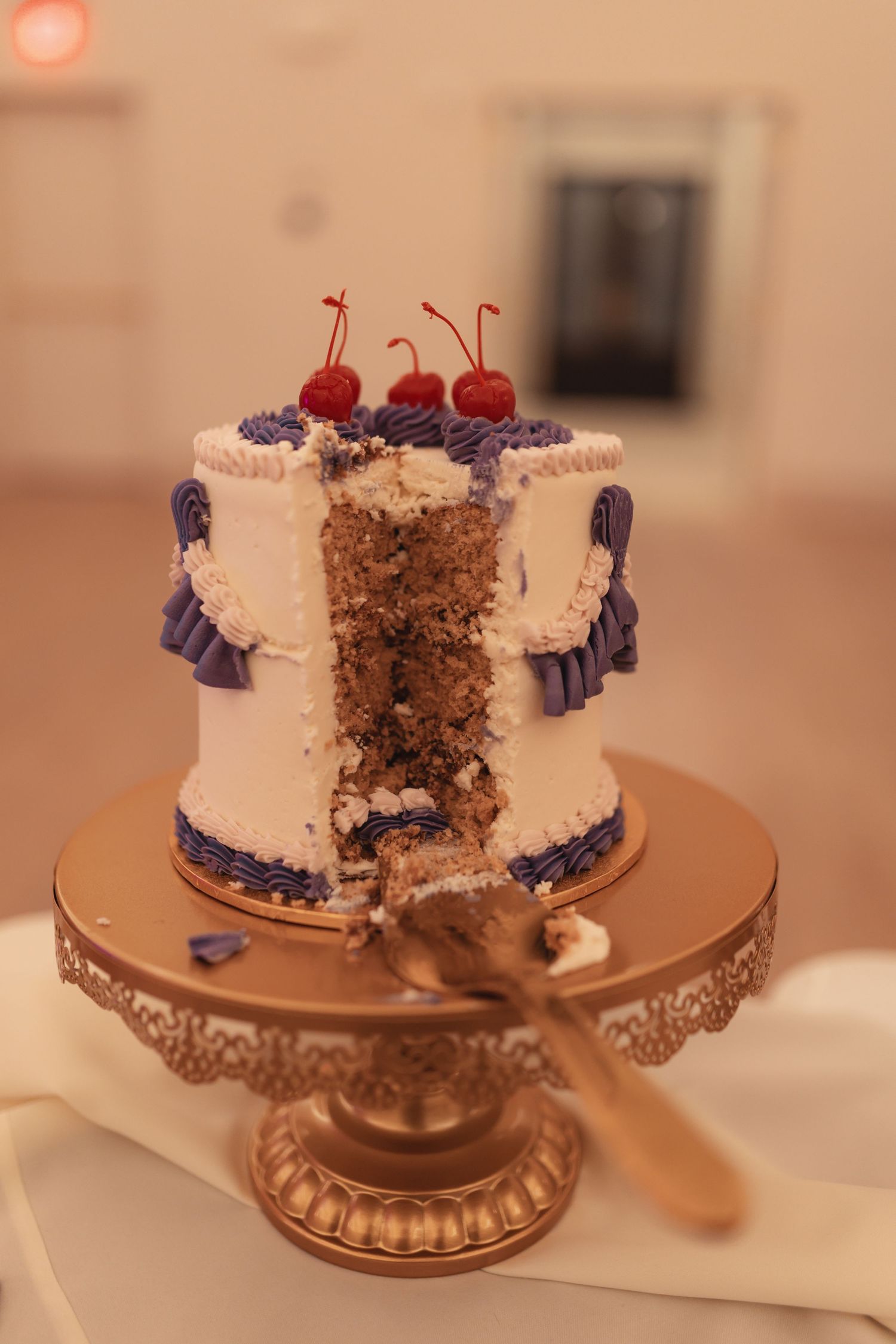 Two-tier white wedding cake with purple frosting and cherries on top, sliced open on a gold cake stand.