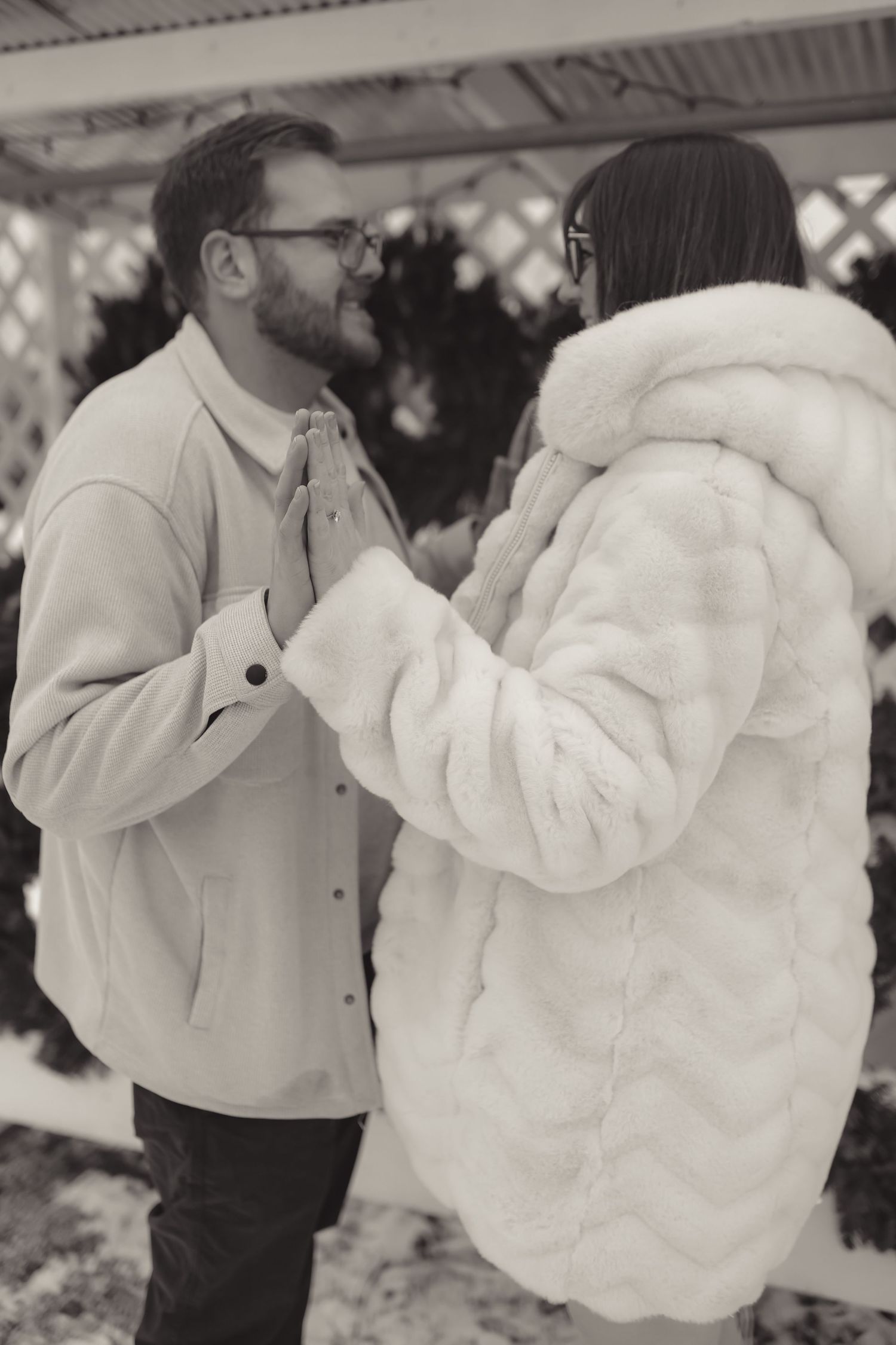 Black and white photo of a couple in winter coats holding hands outdoors near a lattice gazebo in snow.