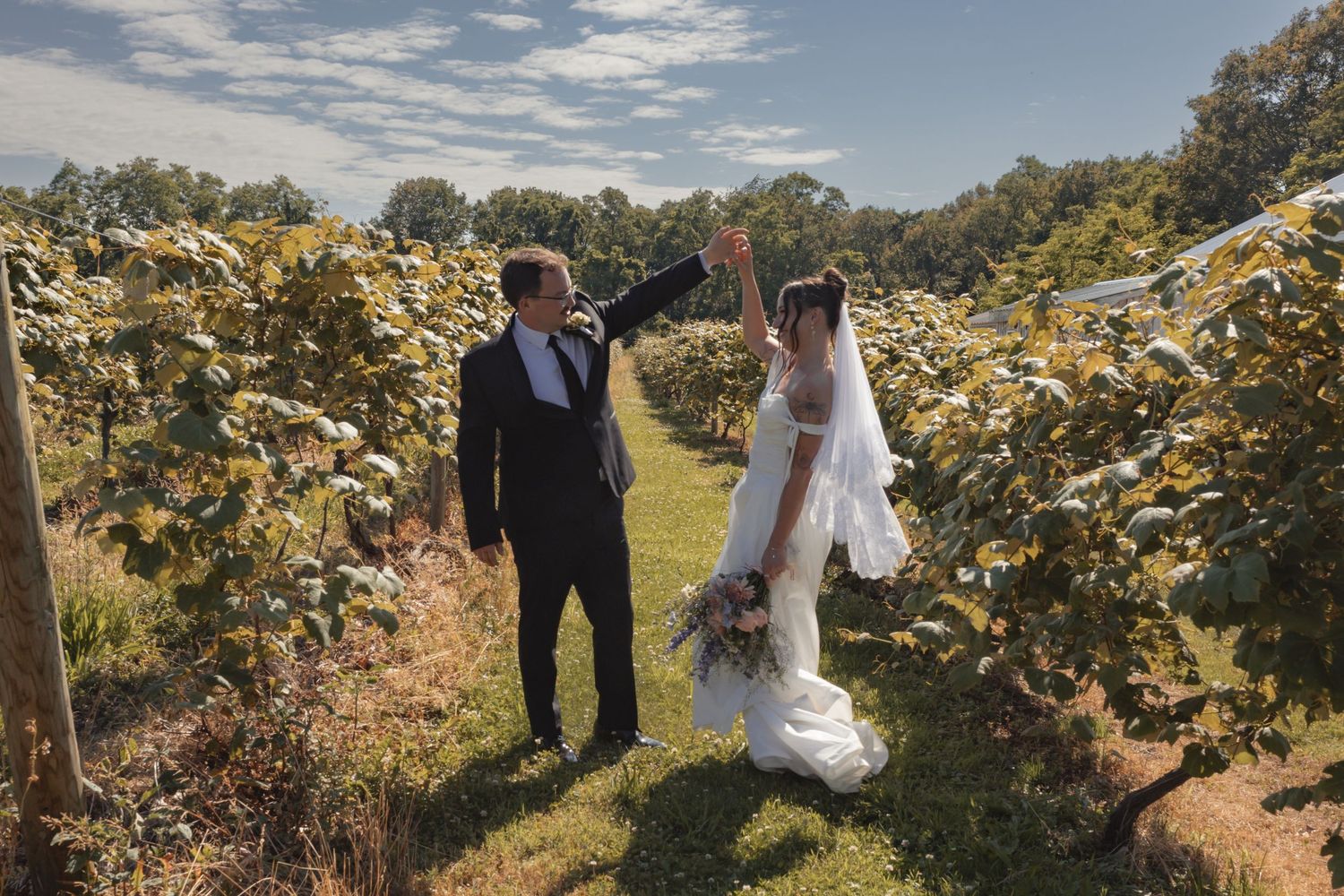 A bride and groom walk hand in hand through a lush vineyard on their wedding day under a bright blue sky.