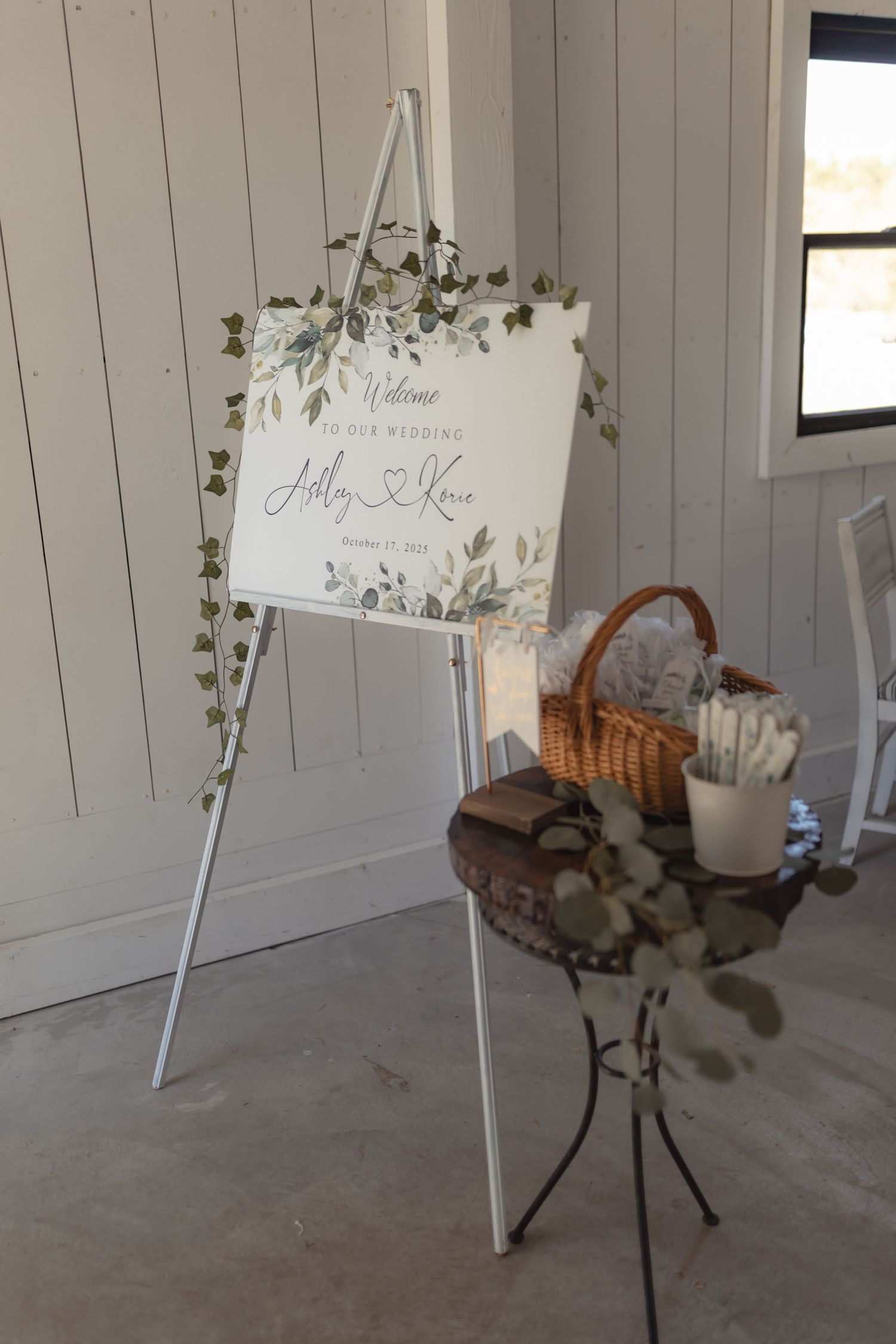 Elegant wedding welcome sign on white easel with greenery decor beside a rustic table with basket.