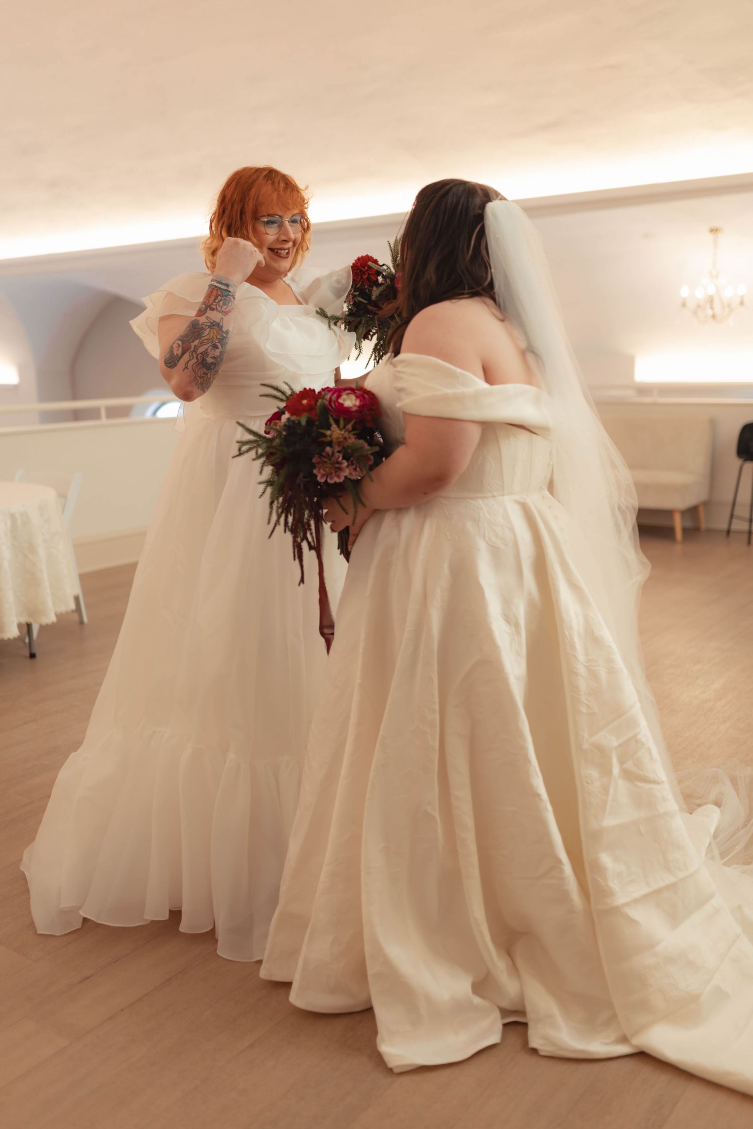 Two brides in white wedding gowns sharing a joyful moment holding bouquets.