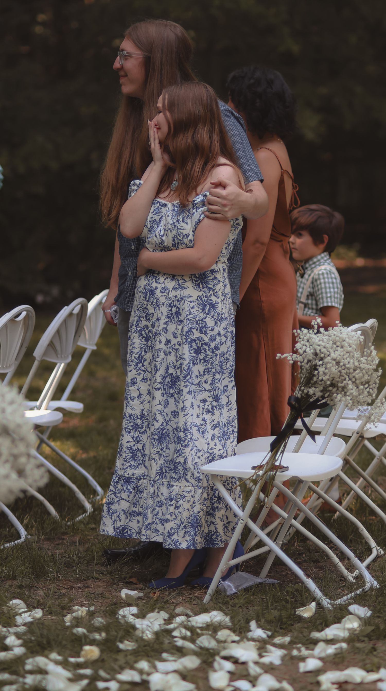 Emotional wedding guests embrace during an outdoor ceremony with white folding chairs and flower petals on the ground.