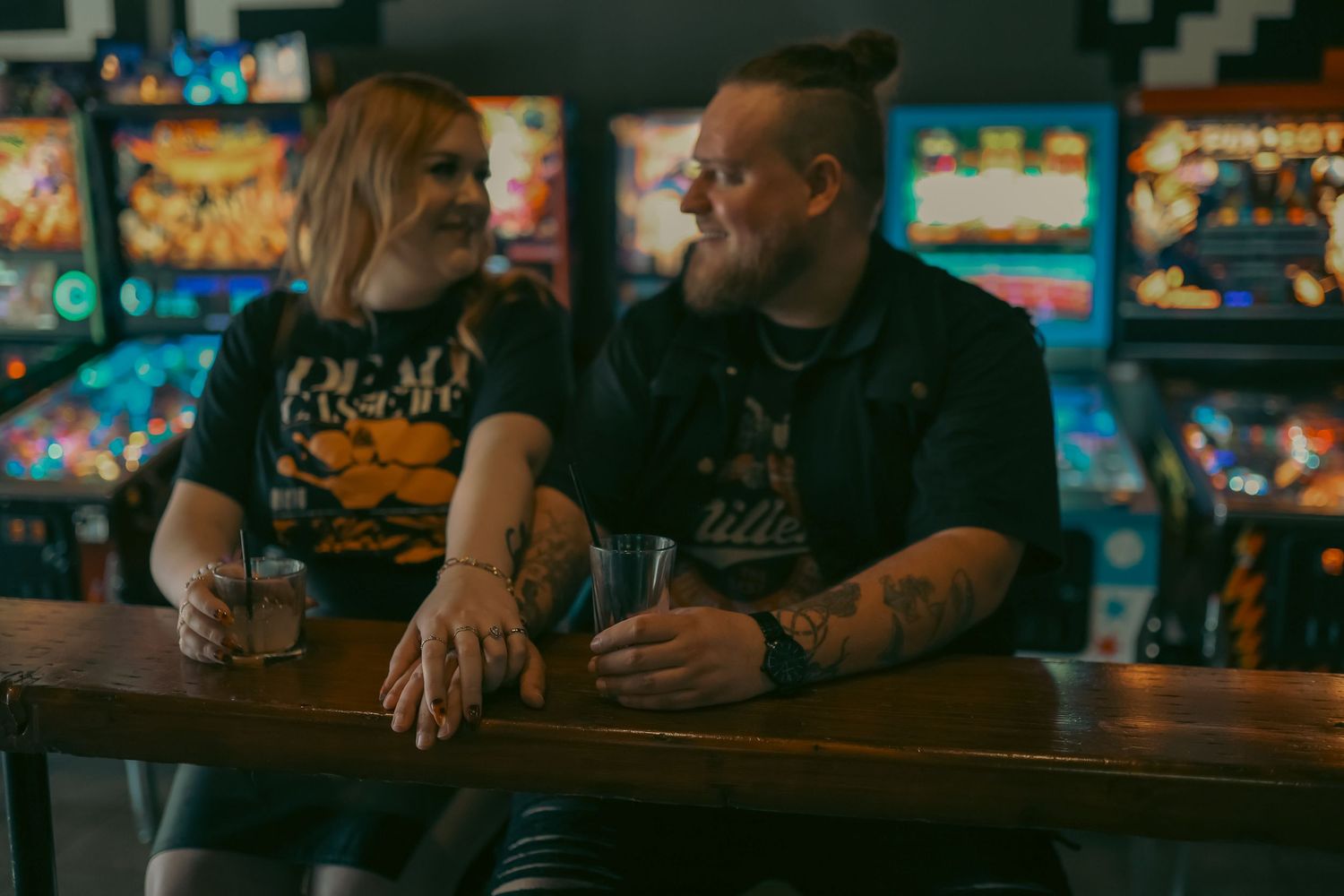 Couple smiling at each other in an arcade bar, holding drinks at a wooden counter with colorful pinball machines behind them.