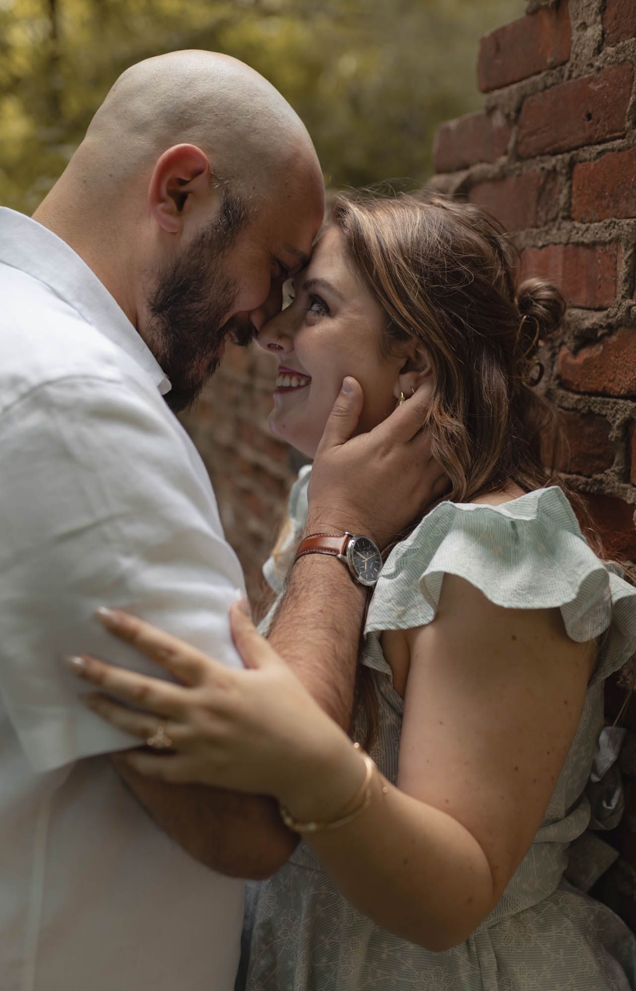 Engaged couple tenderly touching foreheads near a brick wall in a romantic outdoor setting.